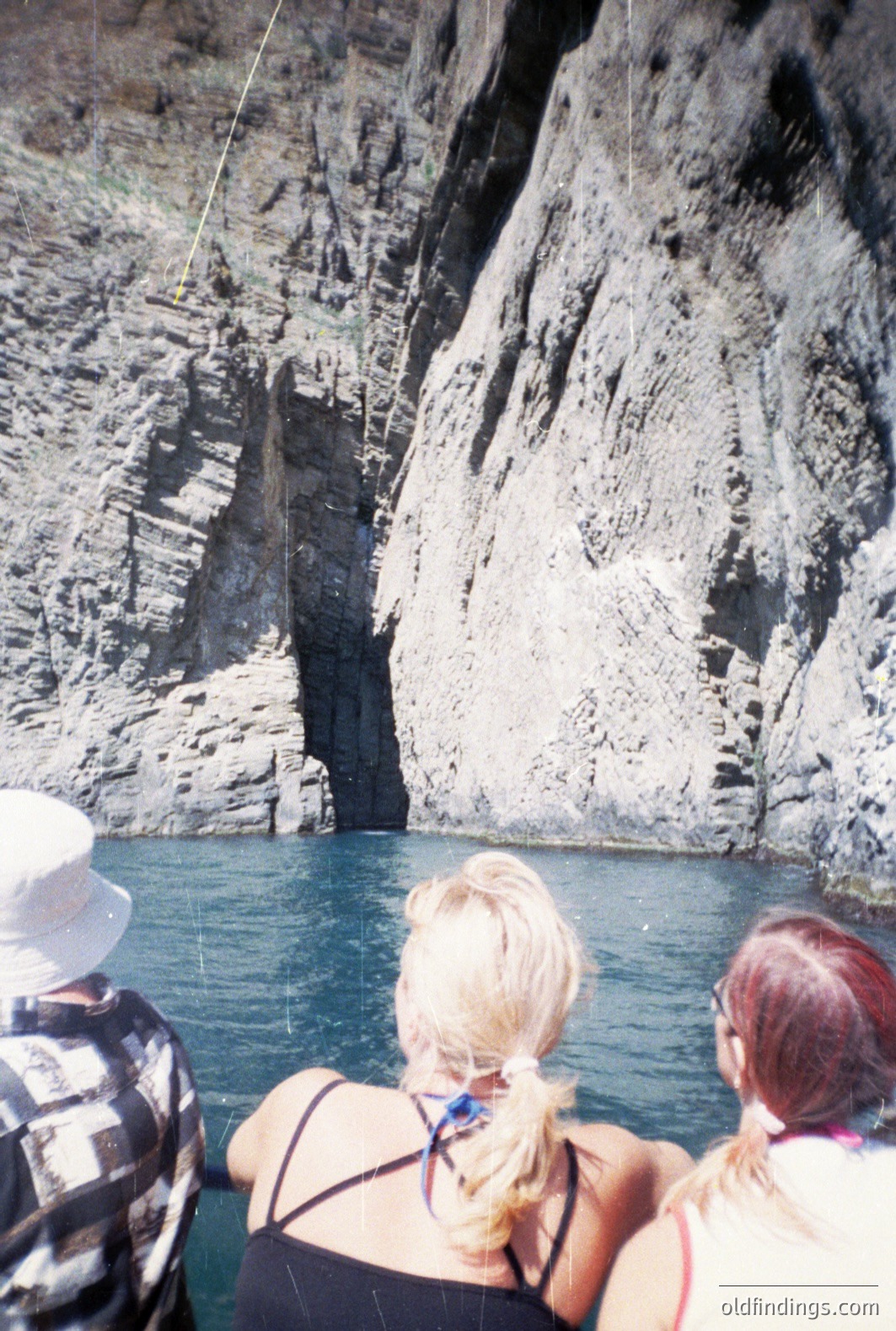A view from a boat, observing towering coastal cliffs with layered rock formations. Two women with blonde and auburn hair are seen from behind. The water is deep blue. Likely a seaside tourist excursion, potentially in the Mediterranean. The photograph's color palette and film grain suggest the 1970s or 80s.