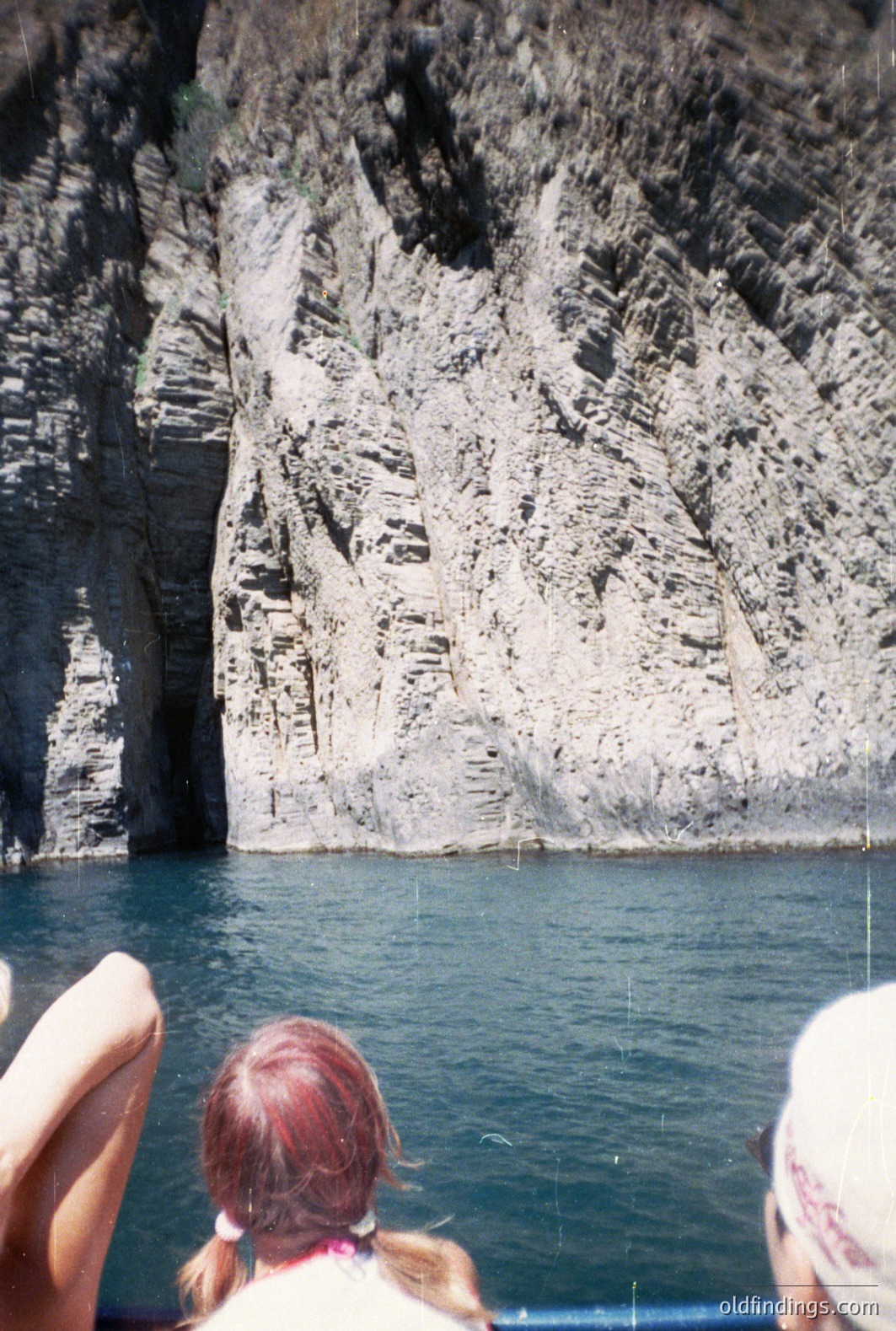 Dramatic coastal cliffs rise steeply from a turquoise sea. The layered sedimentary rock formations exhibit distinct striations and textures. Two figures (partial view) appear on a boat deck, observing the landscape. Likely a tourist photo, possibly 1980s-90s based on film quality. A visually striking natural scene.