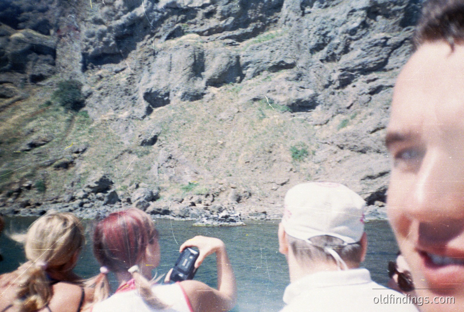 A close-up perspective captures three individuals on a boat tour, framed by the photographer's face. A woman with blonde hair and a red-haired woman are visible, both facing a rugged, grey rock cliff face. The scene suggests a river or lake setting. Appears to be mid-1990s/early 2000s based on clothing and camera.