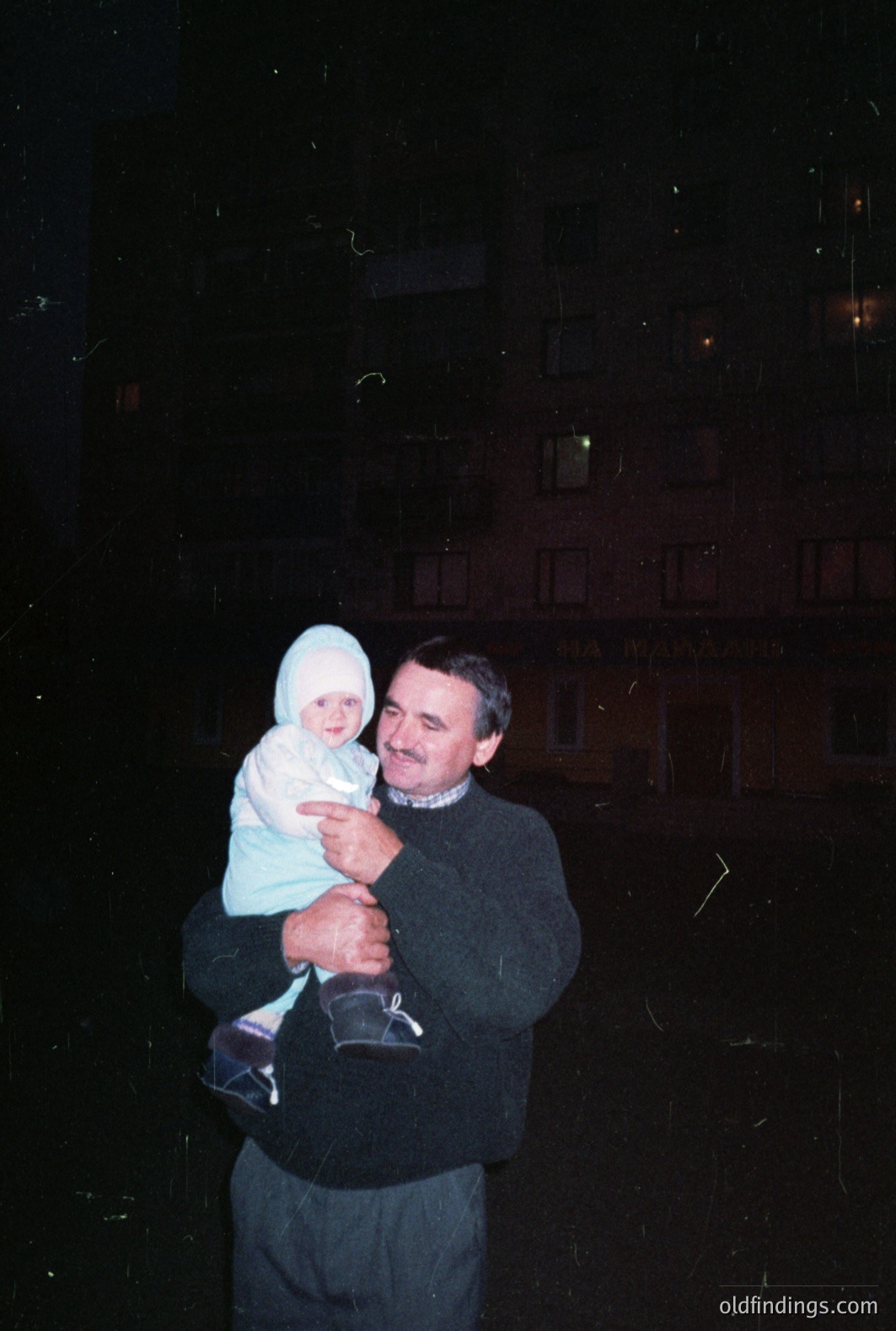 A man carries a baby bundled in a white snowsuit in front of a building with illuminated windows. The building features architectural detailing. Likely a candid portrait, possibly taken in the 1970s or 1980s. The image displays characteristic color and grain. Commercial value: family photography, vintage aesthetics.