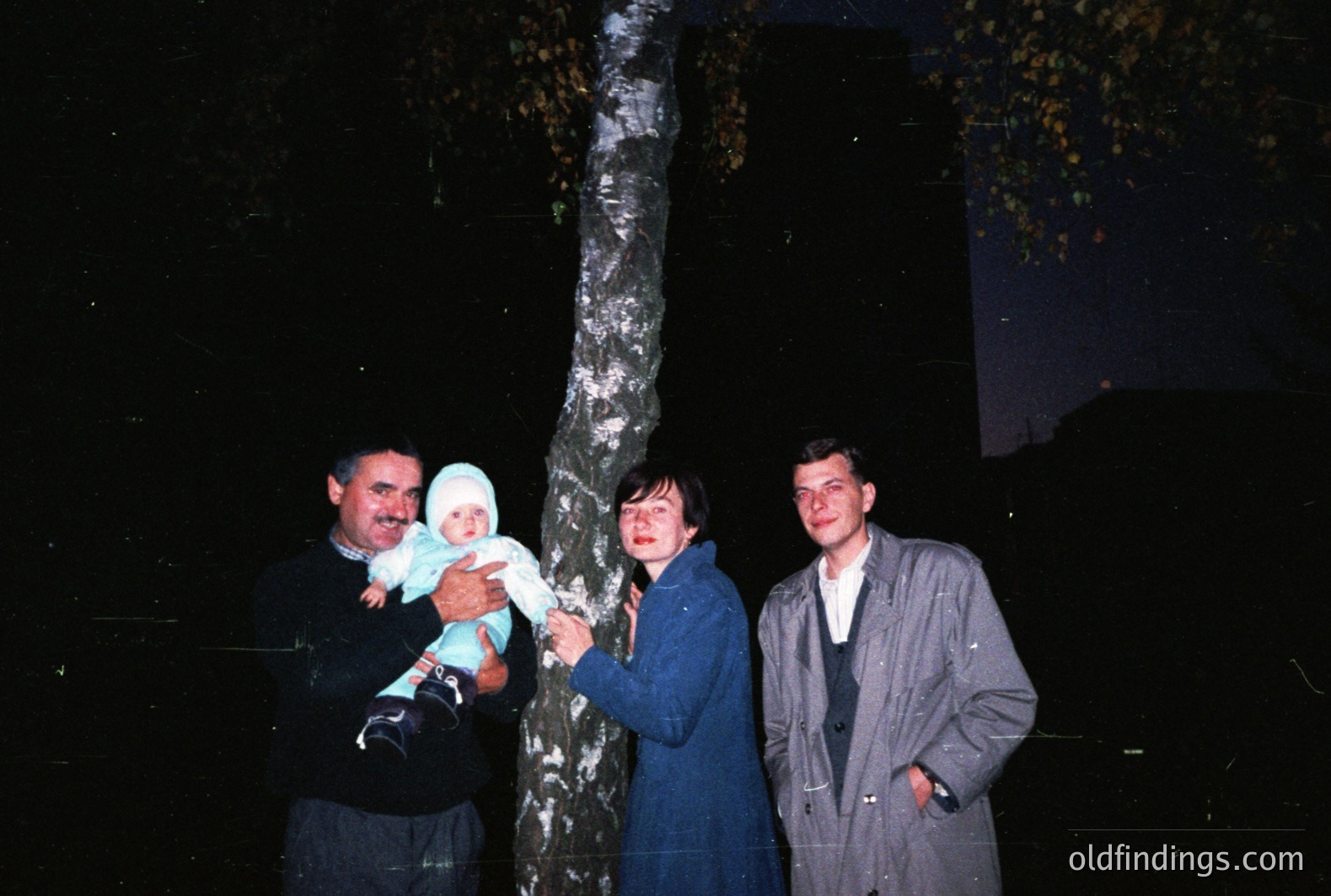 A family portrait, likely a candid snapshot, featuring a man holding a baby, a woman in a blue coat, and a young man, posed near a birch tree. The setting appears to be nighttime, possibly in an Eastern European location. Film grain and color cast are evident, suggesting a 1970s or 1980s origin.