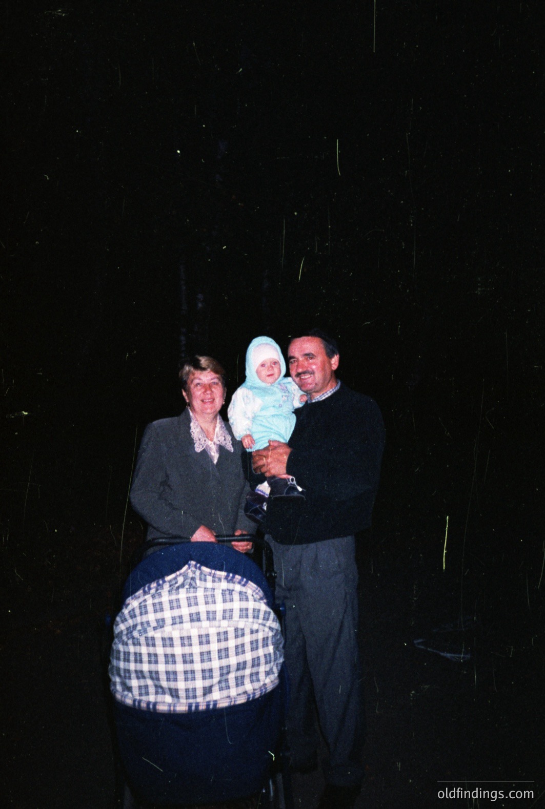 A family portrait: a smiling baby in a white cap is held by a man in a dark sweater, standing next to a woman in a gray plaid suit. Likely 1970s attire. The baby is held in a stroller covered with a dark blue hood. The background is completely black.