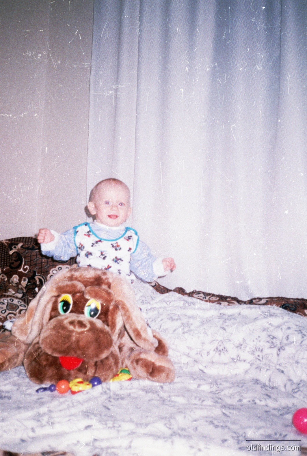 A baby, dressed in a patterned onesie, sits atop a large plush dog toy surrounded by colorful balls. The image appears to be a snapshot, likely taken in a home setting with a plain curtain backdrop. The photograph's color palette suggests a 1980s or early 1990s era. Commercial potential for nostalgic family imagery.