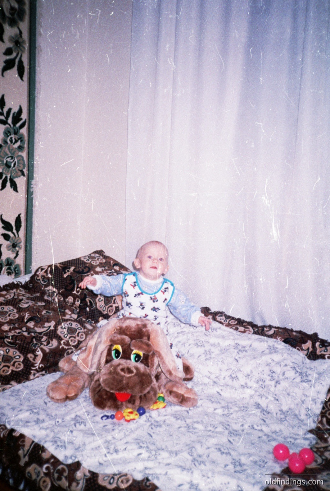 Infant in a patterned onesie sits playfully on a plush, oversized brown dog toy. A patterned bedspread and wallpaper suggest a domestic interior. Pink balloons visible. Likely a family snapshot, circa 1970s-80s. Evokes nostalgia; could be suitable for vintage-themed projects.