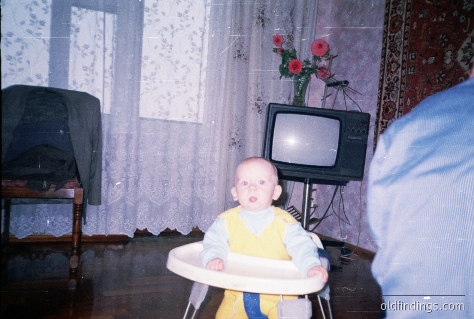 Infant sits in a plastic highchair, gazing directly at the camera. Room features floral curtains, rug, & a vintage television on a metal stand. Likely taken in the 1970s or 80s. Color saturation suggests early color film. Domestic interior scene with strong nostalgic appeal.