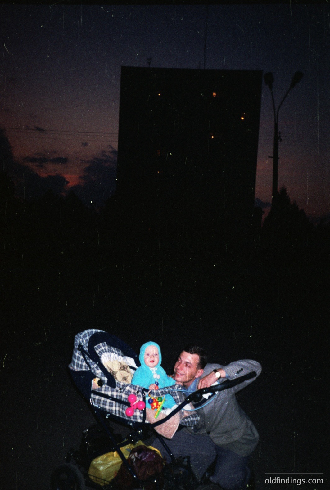 Man pushing stroller with baby in a blue onesie. Stark, Brutalist apartment block dominates the background with lit windows. Appears to be dusk or evening, likely Eastern Europe. Possible 1970s or 1980s aesthetic. Photo exhibits signs of age/film grain. Evokes a nostalgic, residential scene.