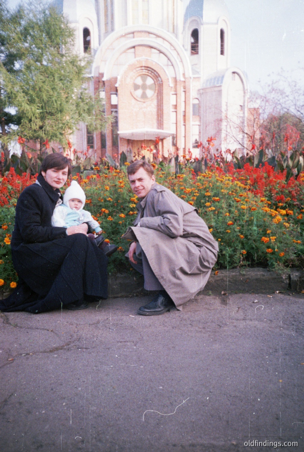 A family of three poses before a grand, ornate Orthodox church building. The mother, in a dark dress, holds a baby wearing a white cap. The father, in a trench coat, squats beside them. Orange flowers bloom in the foreground. Likely Eastern Europe, 1970s.