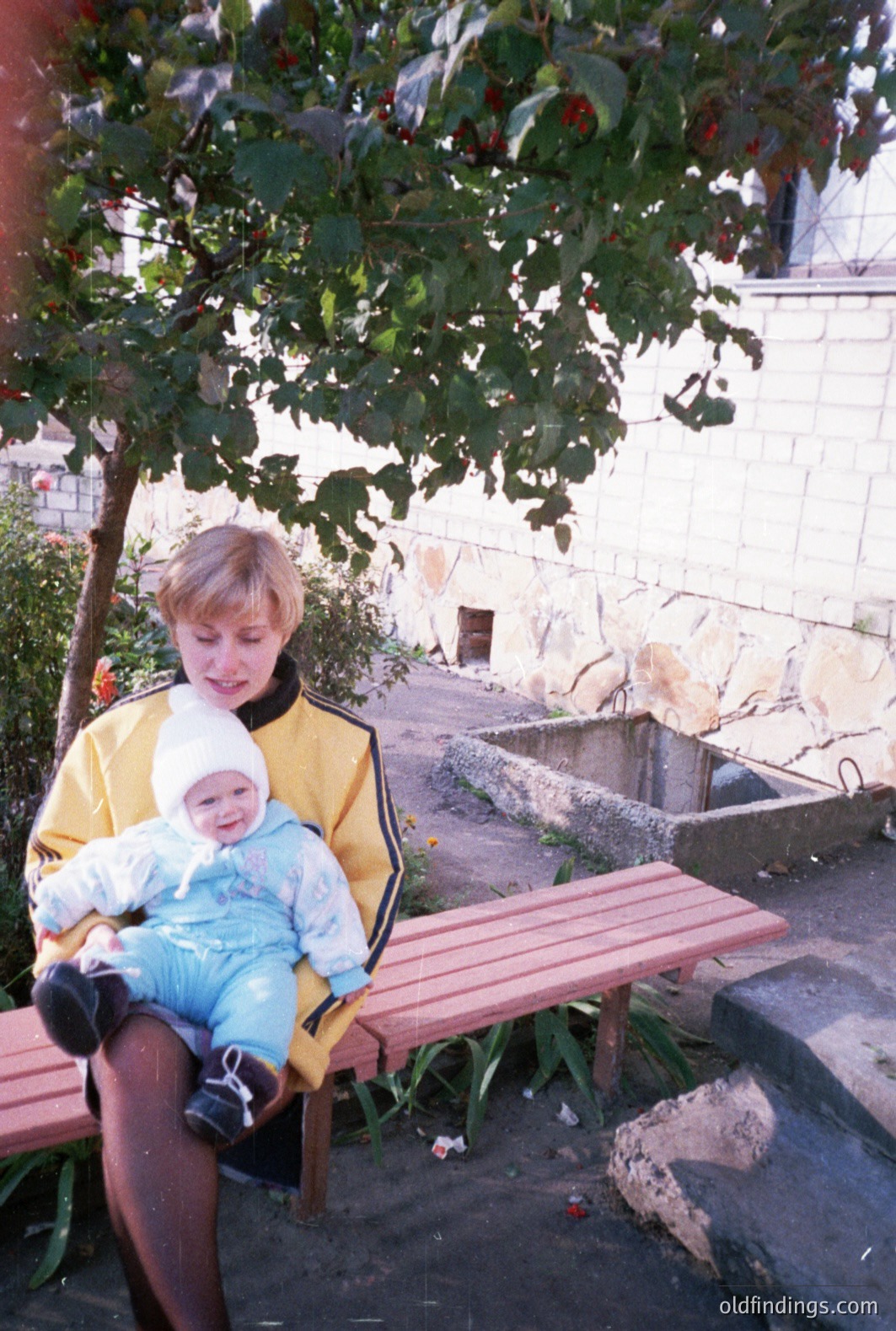 A woman in a yellow track jacket sits on a stone bench, holding a baby wearing a light blue snowsuit. The scene features a small courtyard with a stone wall and foliage. Likely taken in the 1980s or early 90s. The image exhibits characteristics of snapshot photography.