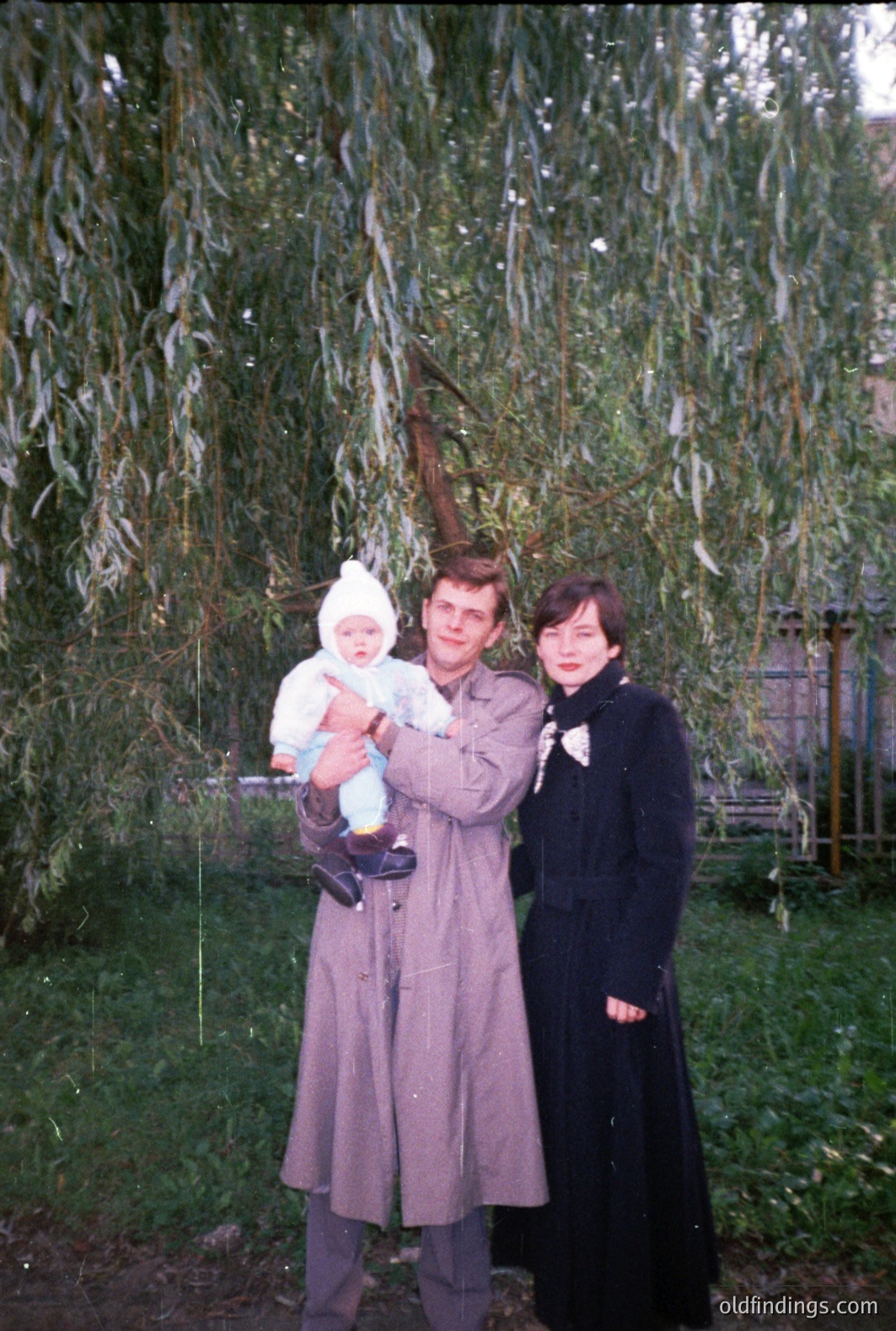 A man in a trench coat holds a baby bundled in white. A woman in a long black coat stands beside them, both facing the camera. A weeping willow tree dominates the background. Appears to be a candid portrait, circa 1970s, potentially Eastern Europe.
