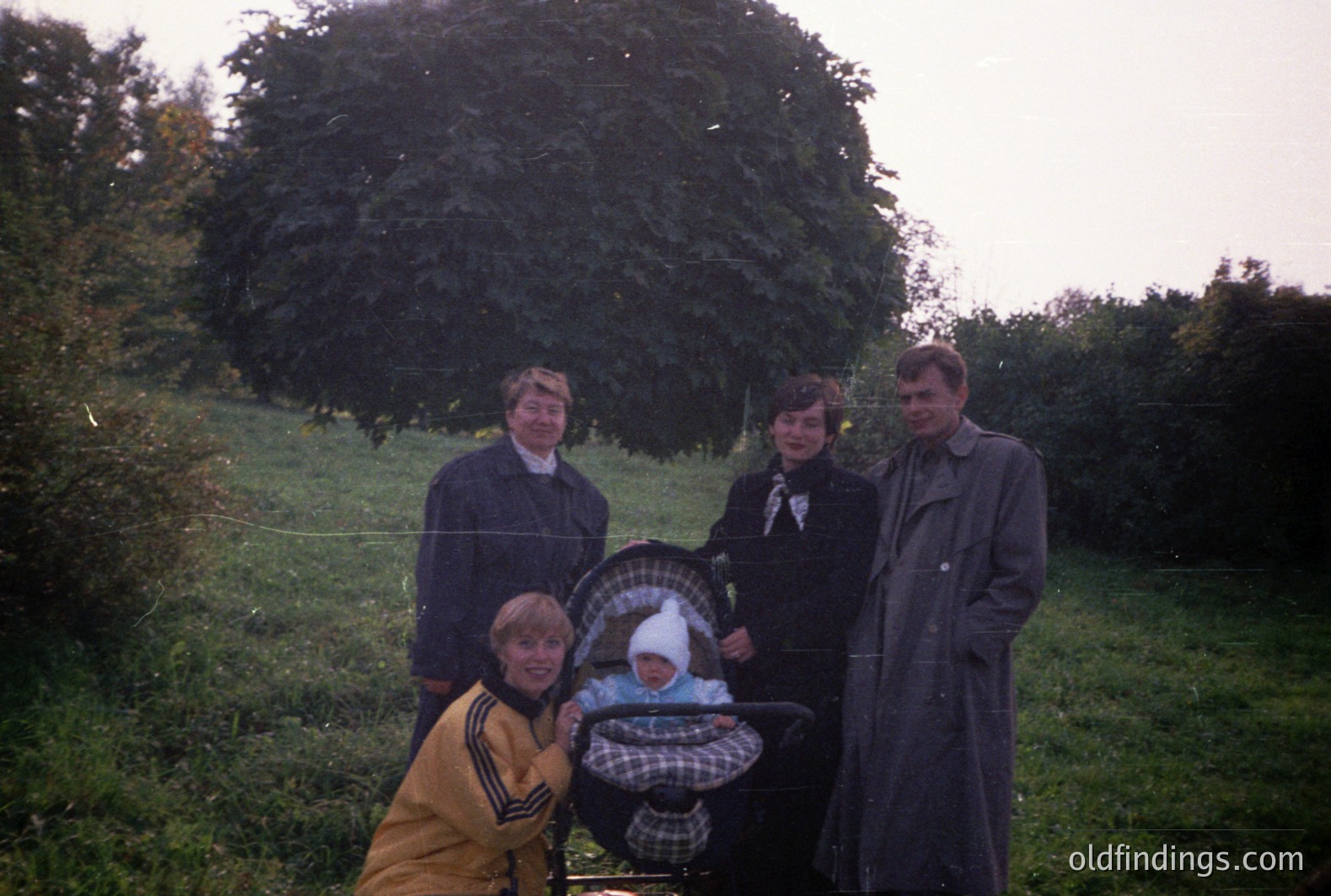 A family portrait, likely taken in the 1970s. Four people pose outdoors near a large evergreen shrub. A baby is in a stroller, viewed partially. The adults wear typical outerwear of the era: trench coats, a track jacket. Grass and foliage fill the background. Simple, candid moment.