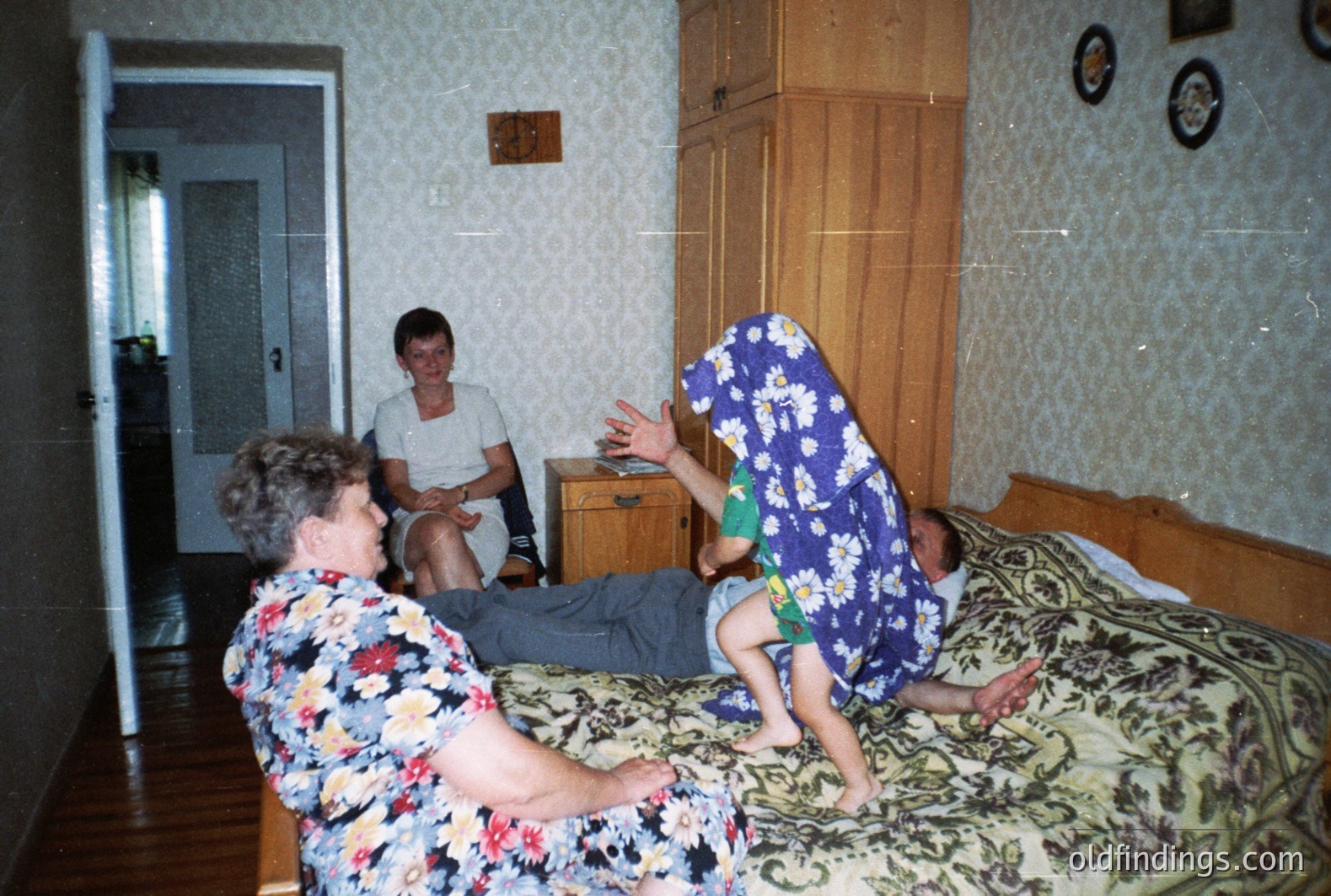 A family scene: A young boy playfully jumps onto a man reclining on a patterned bedspread, while a woman watches with amusement. Interior features 1970s-80s wallpaper, a large wardrobe, and hallway visible. Likely a domestic setting.