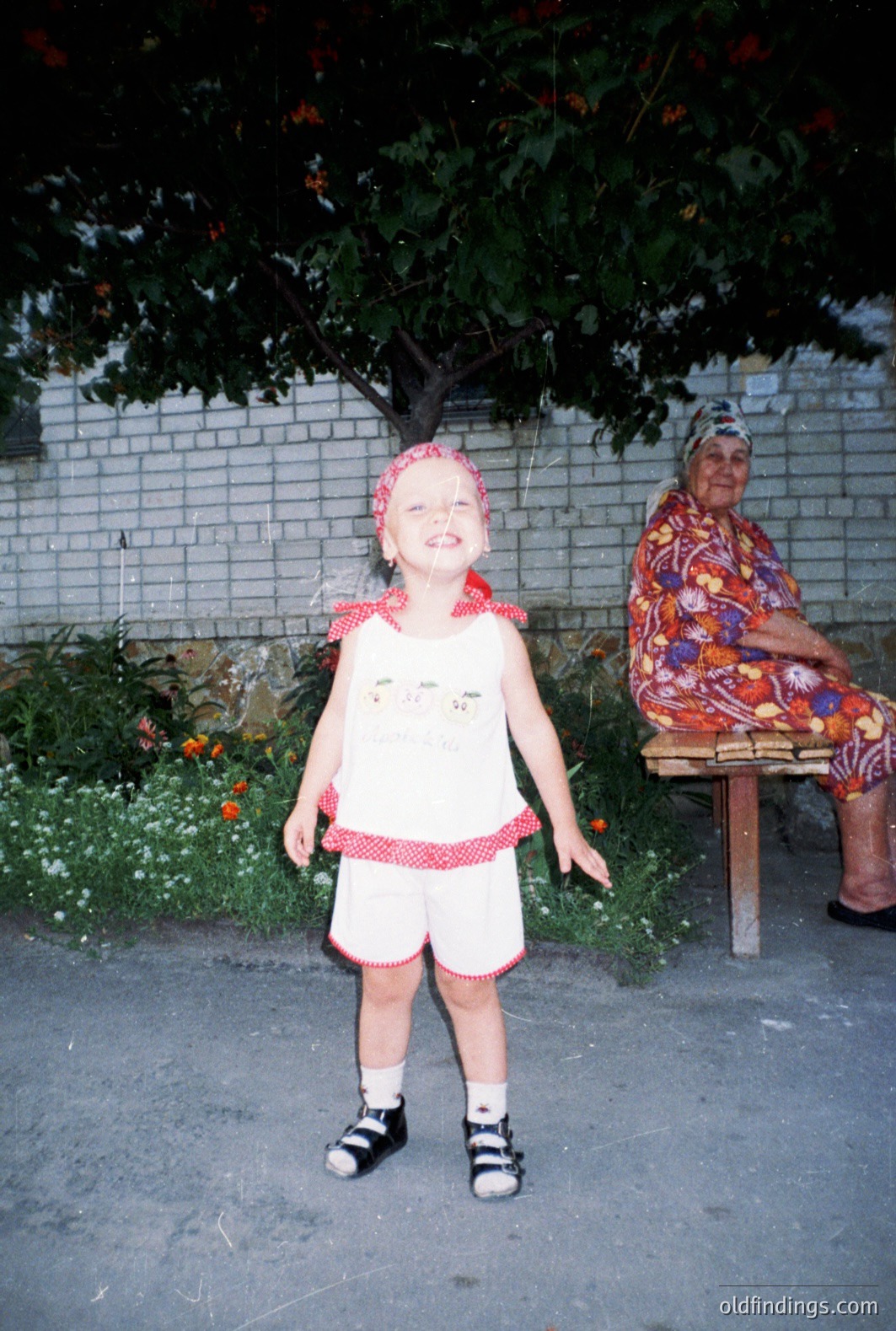 A young child in a white and red patterned dress stands outdoors, facing the camera with a wide grin. An elderly woman sits on a wooden bench nearby. A brick wall and flowering tree form a backdrop. Likely a domestic scene, possibly Bulgaria. Evokes nostalgia; design reference for vintage textiles.