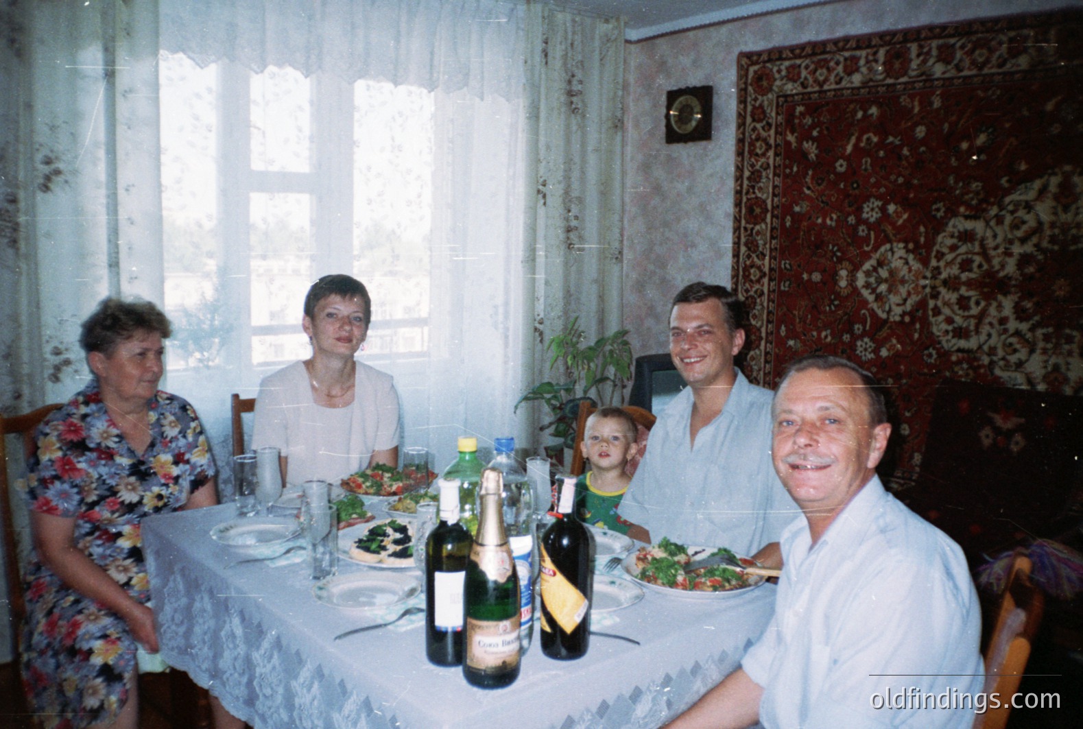 A family gathers around a table laden with food & wine. The setting appears to be a home interior, possibly Eastern European, judging from the decor & clothing styles. Likely a candid snapshot, reflecting a relaxed, casual atmosphere from the 1980s or 1990s.