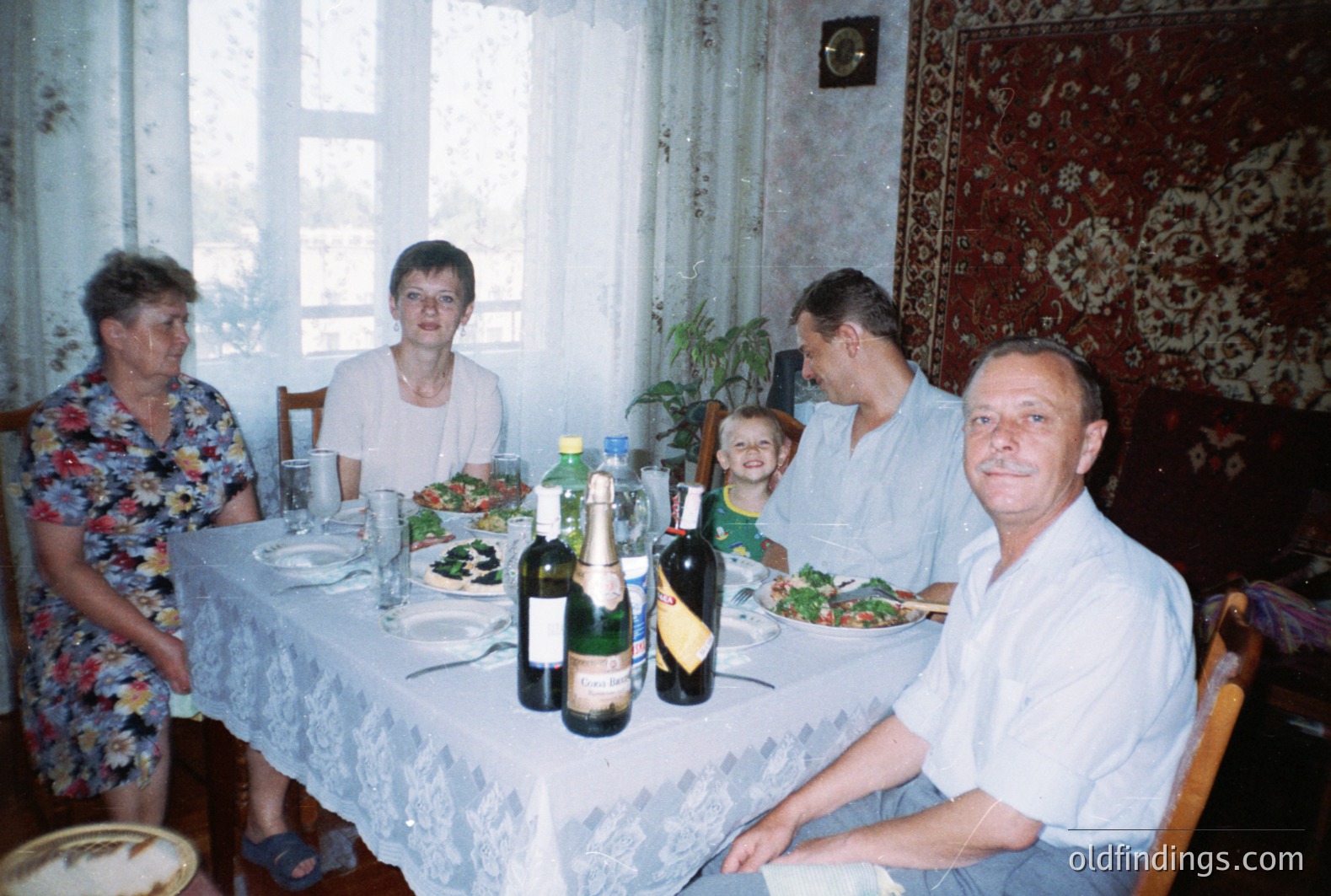A family gathers around a table laden with food and drinks. Visible are several bottles of wine, salads, and bread. The room features a decorative rug, lace tablecloth, and window with floral curtains. Likely a domestic interior, possibly Bulgaria, 1970s-1980s.