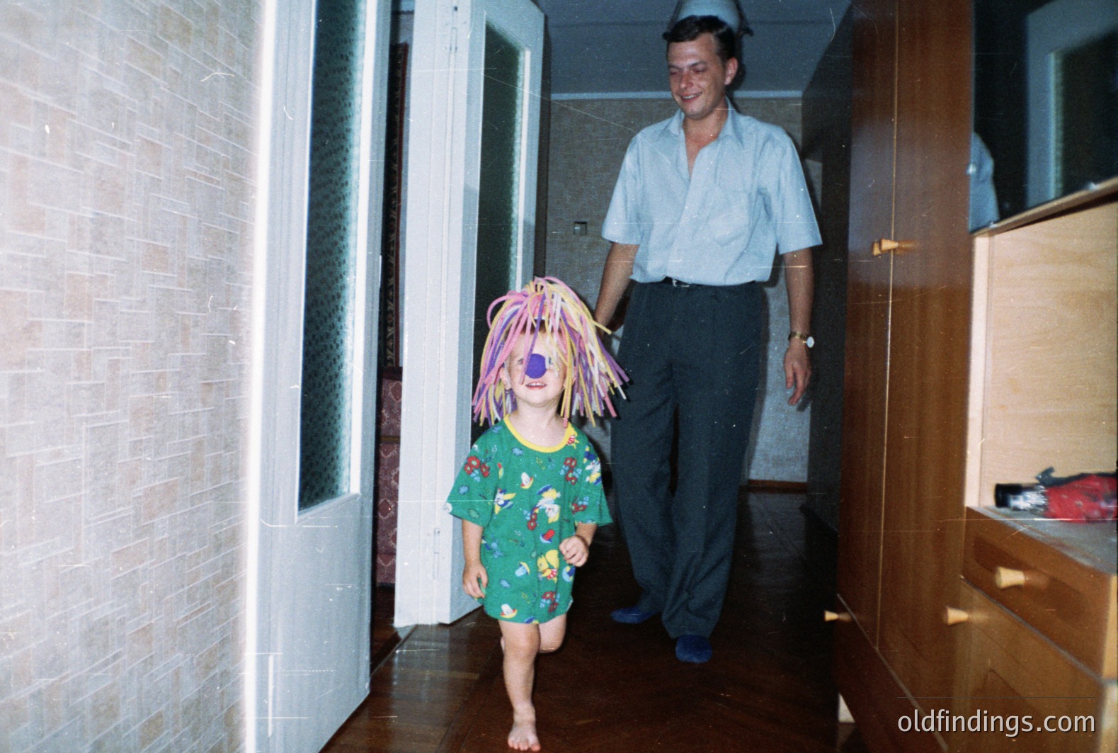 A barefoot child in patterned pajamas and a bright orange wig playfully runs toward the camera in a hallway. An adult man, wearing a blue shirt and dark trousers, walks calmly behind. Likely a 1970s or 80s family snapshot. Interior shows wood floors & built-in wardrobes.