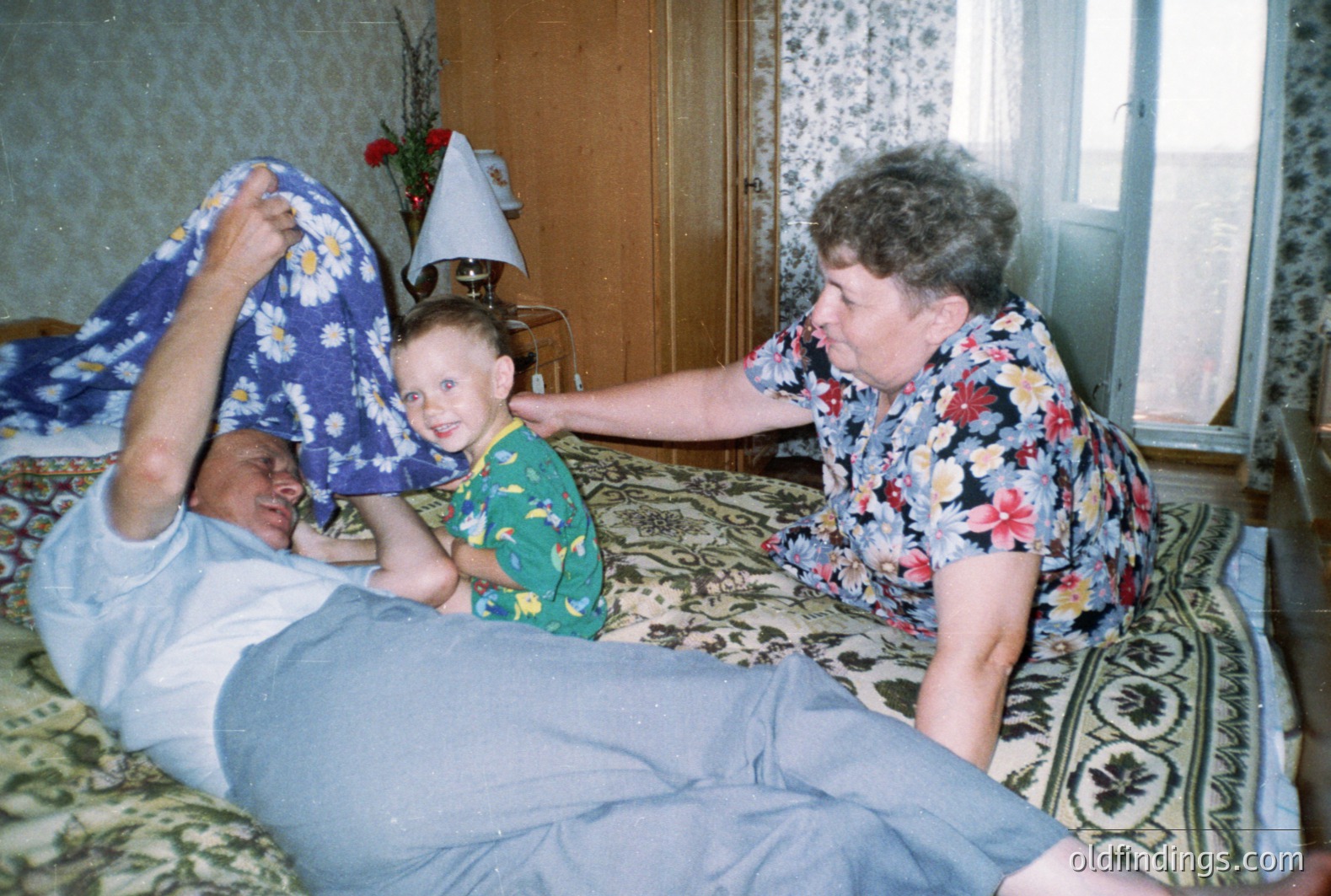 A man lies in bed, playfully raising a floral-patterned blanket while a young boy, in a green shirt, stands nearby. An older woman, wearing a vibrant floral shirt, stands close by, appearing to interact with the scene. Interior view; likely a family moment. Appears to be a 1980s snapshot.