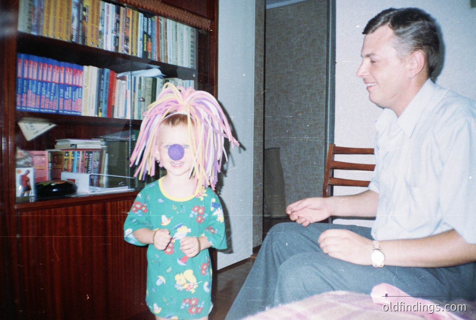 A young child wearing a bright pink and green textured wig stands playfully, covered by one eye patch. A man in a button-down shirt sits nearby, smiling. A large bookshelf filled with books and a record player dominate the background. Likely a snapshot from the 1970s or 1980s.