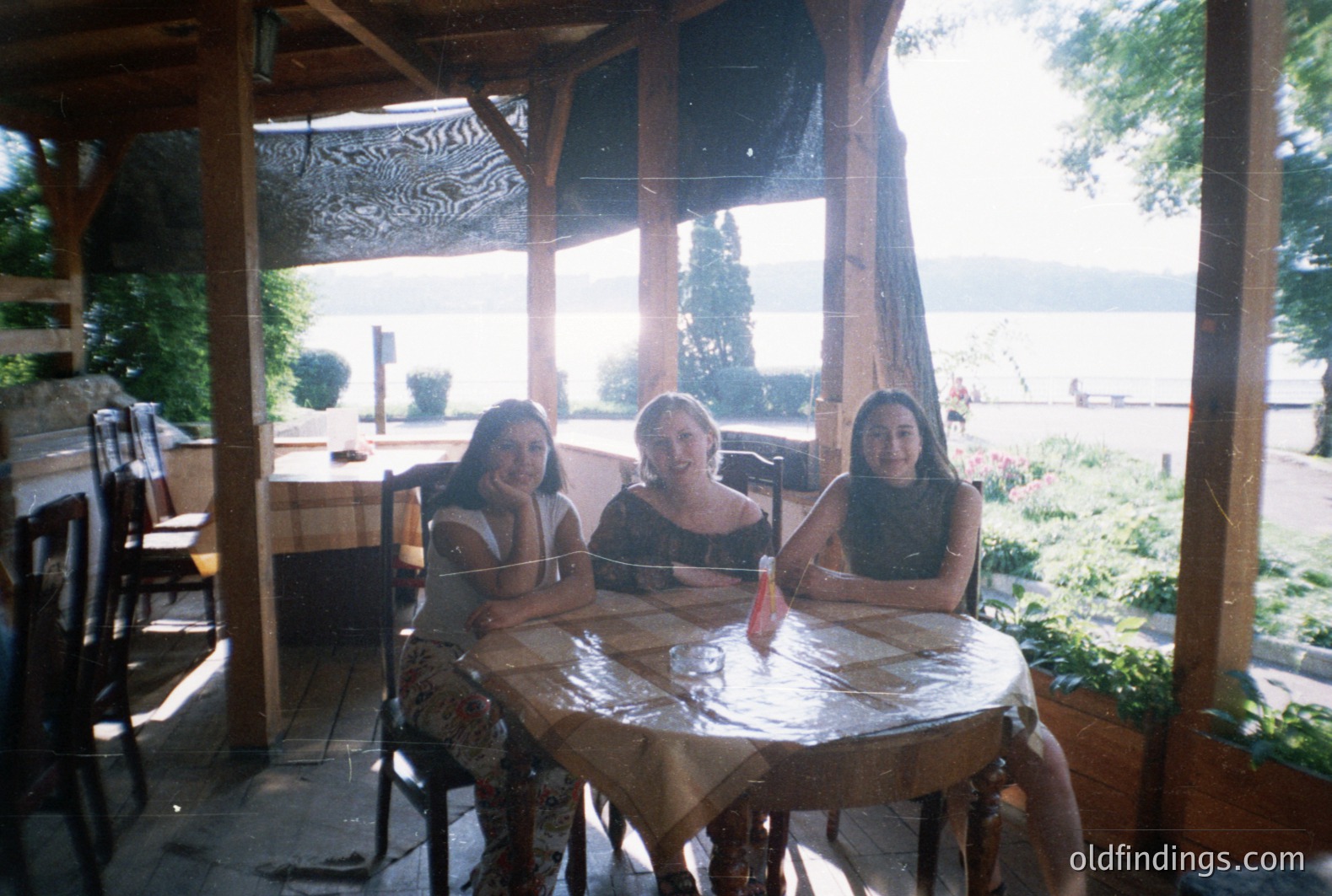 Three young women seated around a table covered with a floral tablecloth, framed by a wooden pergola. A scenic lakeside view is visible through the structure. The photo’s warm tones suggest a 1980s or 1990s snapshot. Likely a resort or recreational area.