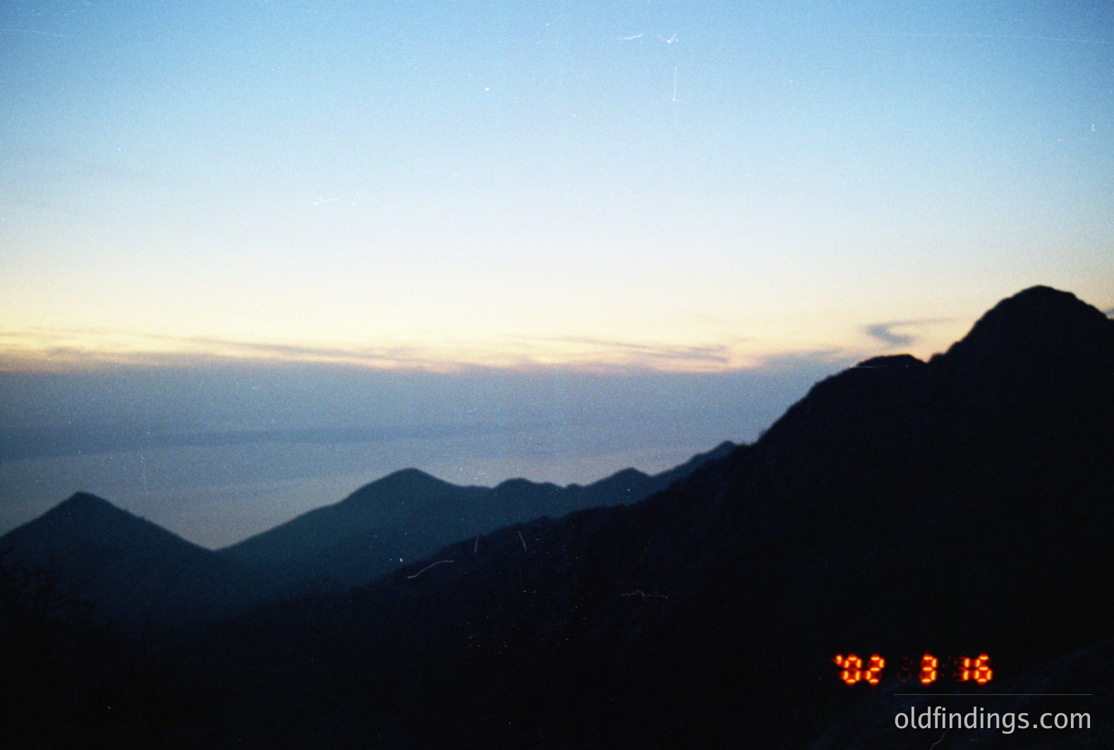 Dramatic, high-altitude landscape at twilight. Silhouetted mountain ridges descend towards a hazy, reflective body of water. A timestamp '02:31:16' is visible in the corner, likely from the camera's internal clock. Likely taken in the 1990s or 2000s. Stock potential for background scenery or environmental themes.