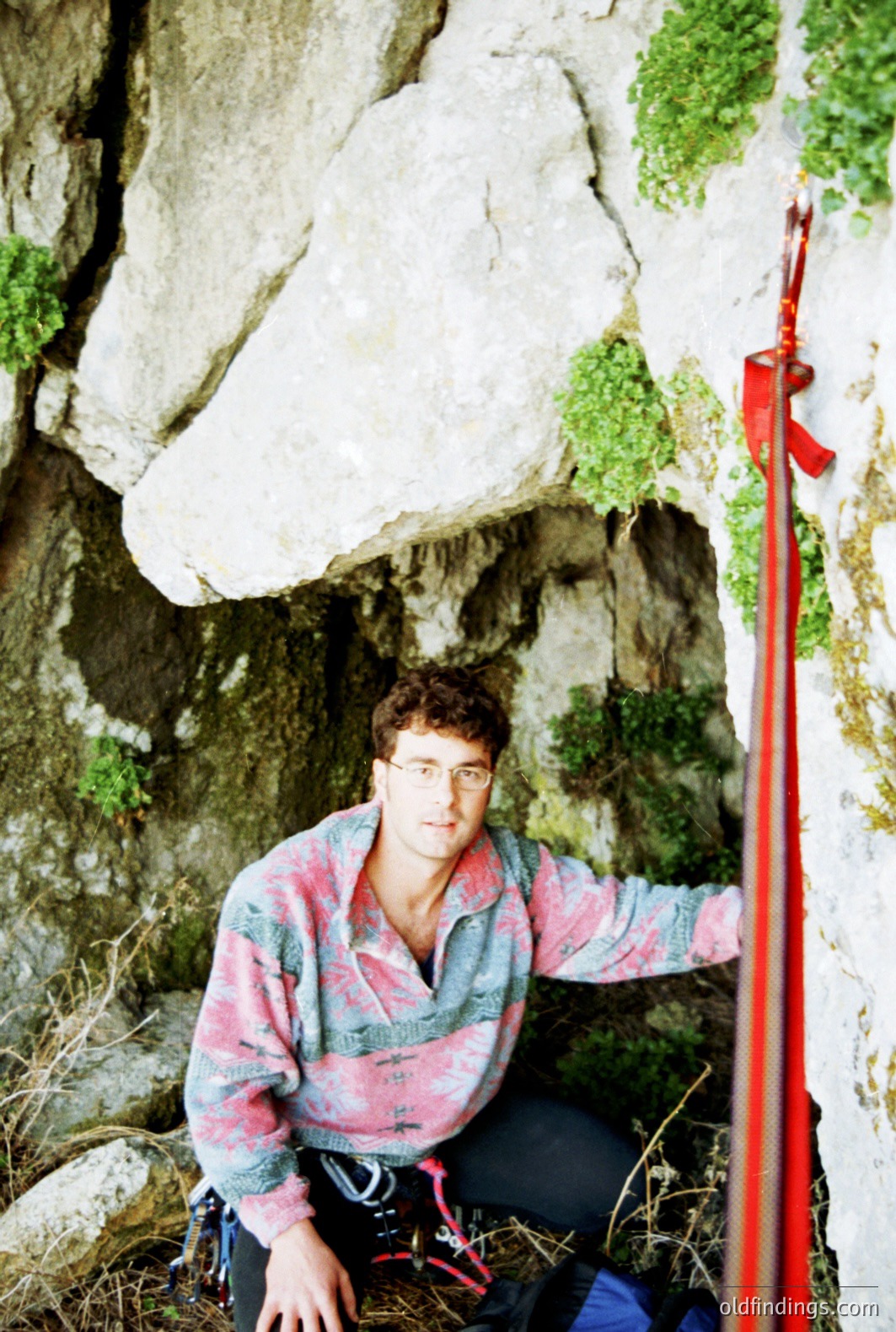 A climber, wearing glasses and a patterned fleece, poses within a rocky overhang. Equipped with climbing gear including ropes and harnesses, he appears to be resting during a climb. Likely a promotional or instructional climbing image, likely 1980s or 90s.