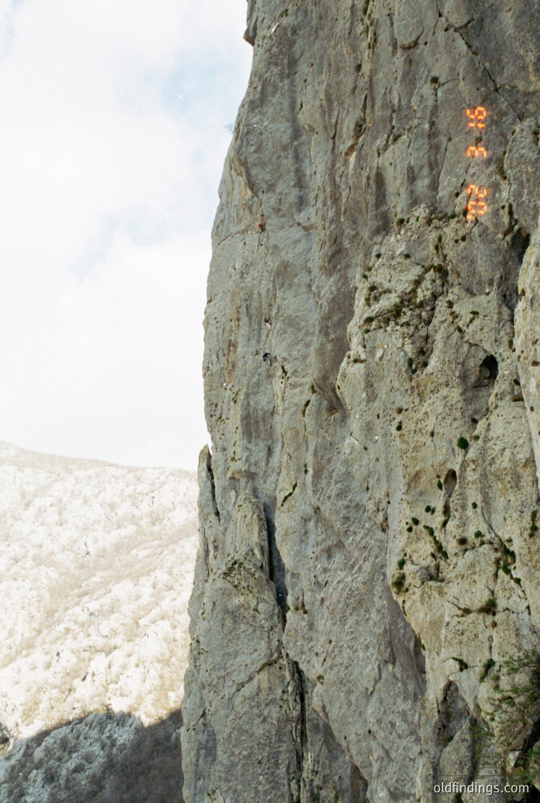 A lone rock climber ascends a sheer, grey cliff face, likely in an alpine setting. Snow-covered peaks are visible in the background. The image shows evidence of a date stamp, "02-16". Potential stock photo use for outdoor adventure, climbing gear, or landscape design.