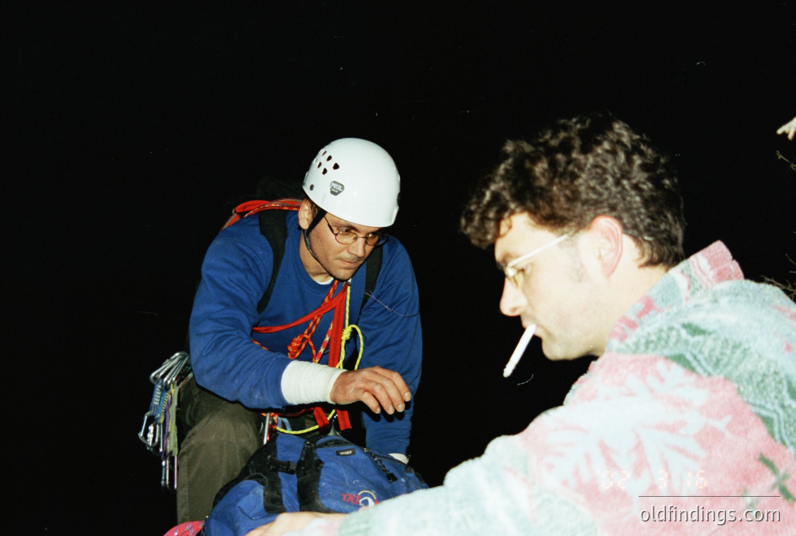 Two men, one wearing a climbing helmet, harness, and backpack, appear to be resting during a climbing activity. One man is smoking a cigarette. The image’s dark background suggests a nighttime setting. Likely taken in the 1990s due to clothing & photographic style.