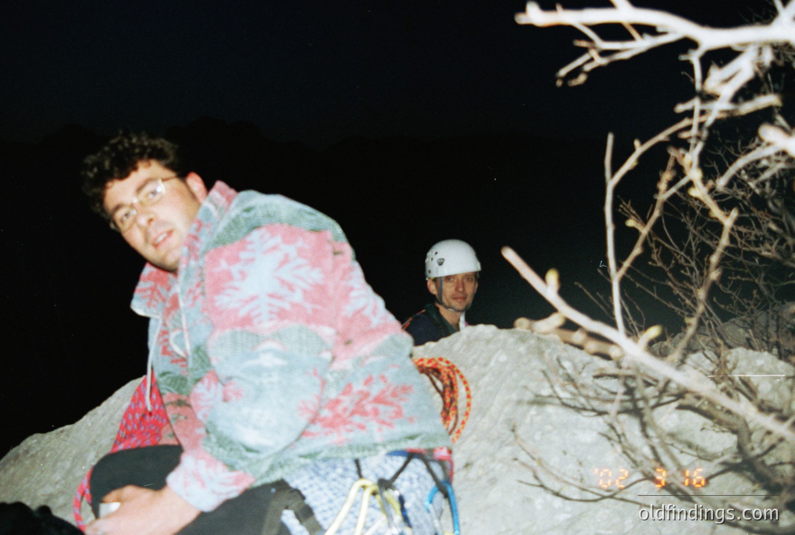 A climber, wearing a patterned puffer jacket and glasses, pauses on a rocky cliff face. Another climber is visible behind, wearing a helmet. Marked "02-23-96" date in the lower right corner suggests mid-1990s. Likely an alpine or mountainous setting. Stock potential for adventure/outdoor themes.