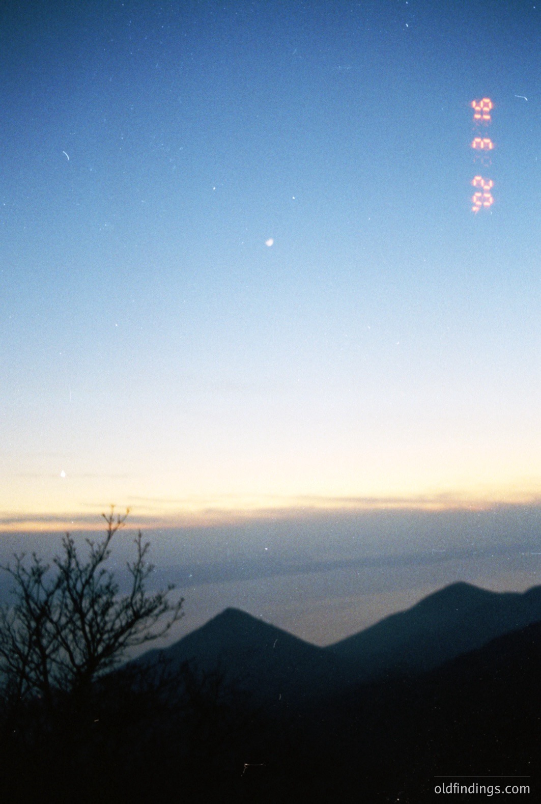 Dramatic twilight landscape with silhouetted mountains, visible sea, and a vibrant orange/yellow horizon. Sparse tree branches in foreground. Appears to be a low-light, possibly nighttime, photograph with a dated film aesthetic. Likely coastal region.