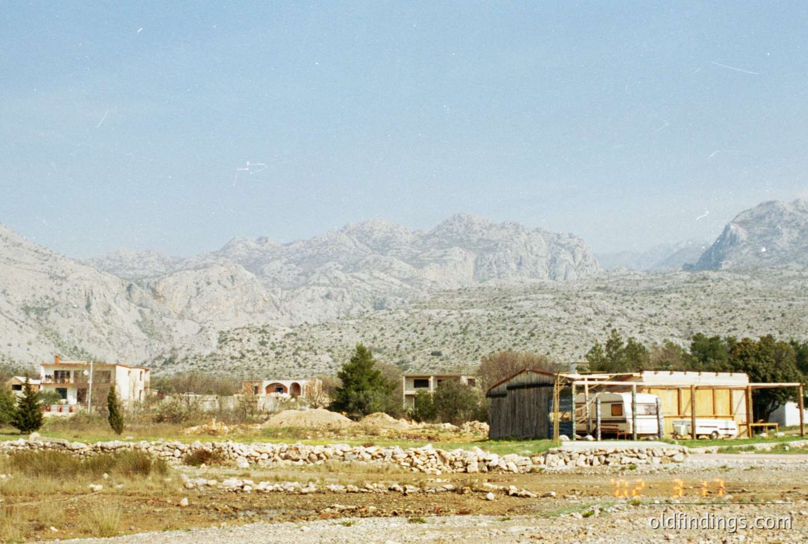 A view of rural buildings and a roadside structure set against a backdrop of dramatic, rocky mountains. Appears to be a transitional landscape, potentially showing early development. Likely Southern Europe, mid-20th century. Rustic charm suggests potential for travel/tourism stock.