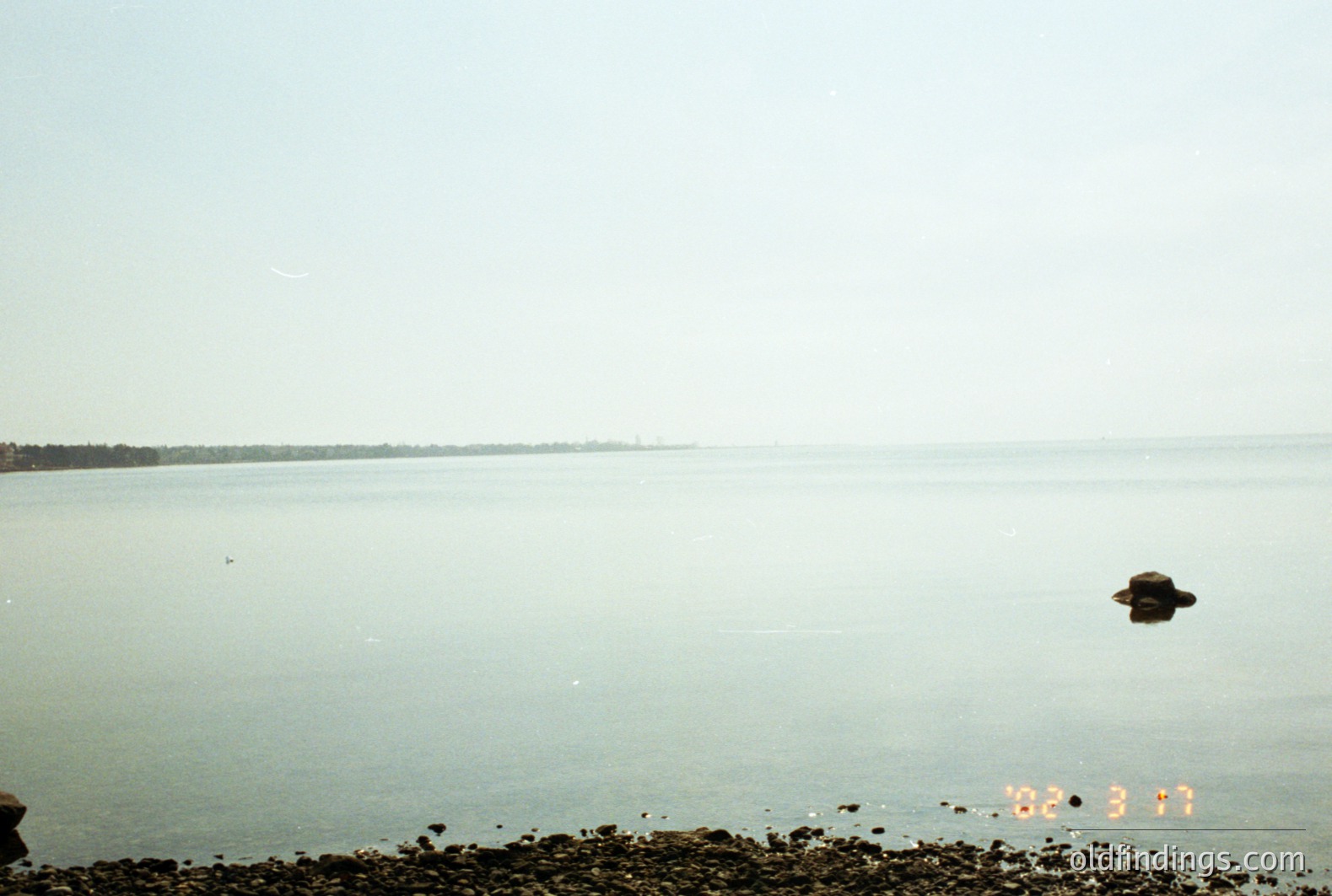 Ethereal seaside scene depicts a calm, hazy body of water stretching to a distant treeline. A rocky foreground adds texture to the shoreline. The image’s soft focus and slightly faded colors suggest a 1970s or 80s film photograph. Potential commercial value for nature stock or design.
