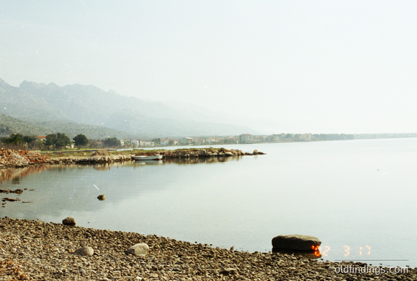 A coastal scene featuring a calm body of water, possibly the Black Sea, framed by a rocky shoreline and distant, hazy mountains. A small boat rests near the bank; a residential area is visible across the water. The photo appears to be a snapshot from the 1990s or 2000s given the film quality and date stamp. Likely Bulgaria, near Varna.