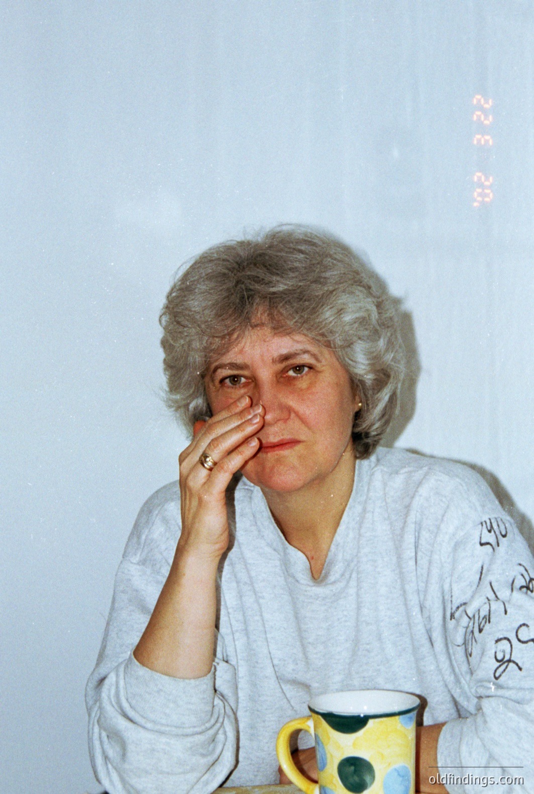 Close-up portrait of a woman with a stylish, layered haircut, seated and appearing thoughtful. She wears a gray sweatshirt & a ring on her finger. A colorful mug sits on a nearby table. Image date stamp visible: 8/28/00. Likely casual snapshot.