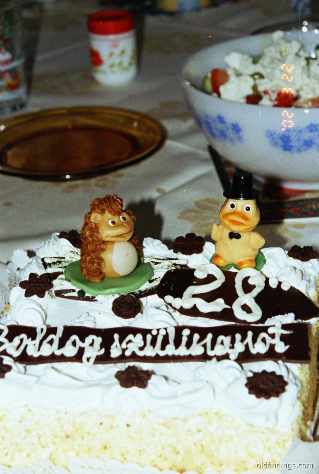 A layered, elaborate celebration cake featuring figurines of a hedgehog and a penguin in a bow tie. Black lettering spells "Boldog szülinapot" (Happy Birthday). The cake’s décor includes chocolate swirls, buttercream frosting, and what appears to be edible flowers. Likely a 1980s or early 90s home snapshot.