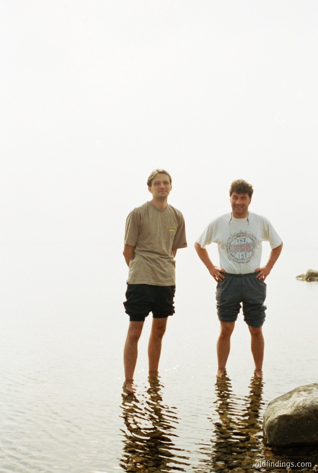 Two young men stand in shallow water, chest-deep, facing the camera. They wear casual summer attire: t-shirts and shorts. Gentle ripples distort reflections. The image evokes a relaxed, possibly lakeside or coastal scene. Likely mid-1990s, judging from clothing styles. Potential stock image value.