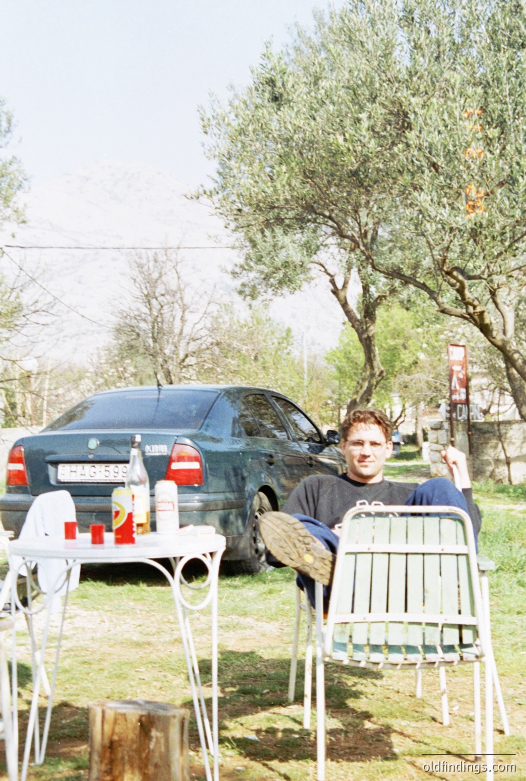 Man seated outdoors, appearing relaxed in a metal chair, near a dark green 1990s Mercedes sedan. The scene features a rustic, slightly overgrown backyard with a glimpse of distant buildings. Likely late 1990s, possibly Eastern European setting based on car registration. Evokes a casual, domestic moment.