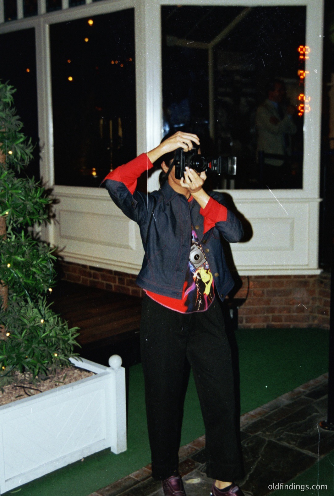 A person in a vibrant, patterned sweater and black pants holds a vintage camera, seemingly taking a photo of the brightly lit exterior. The scene features a decorated evergreen tree, a stone-paved area, and a glimpse of a building with visible signage. Likely captured in the 1990s, exhibiting a snapshot aesthetic.