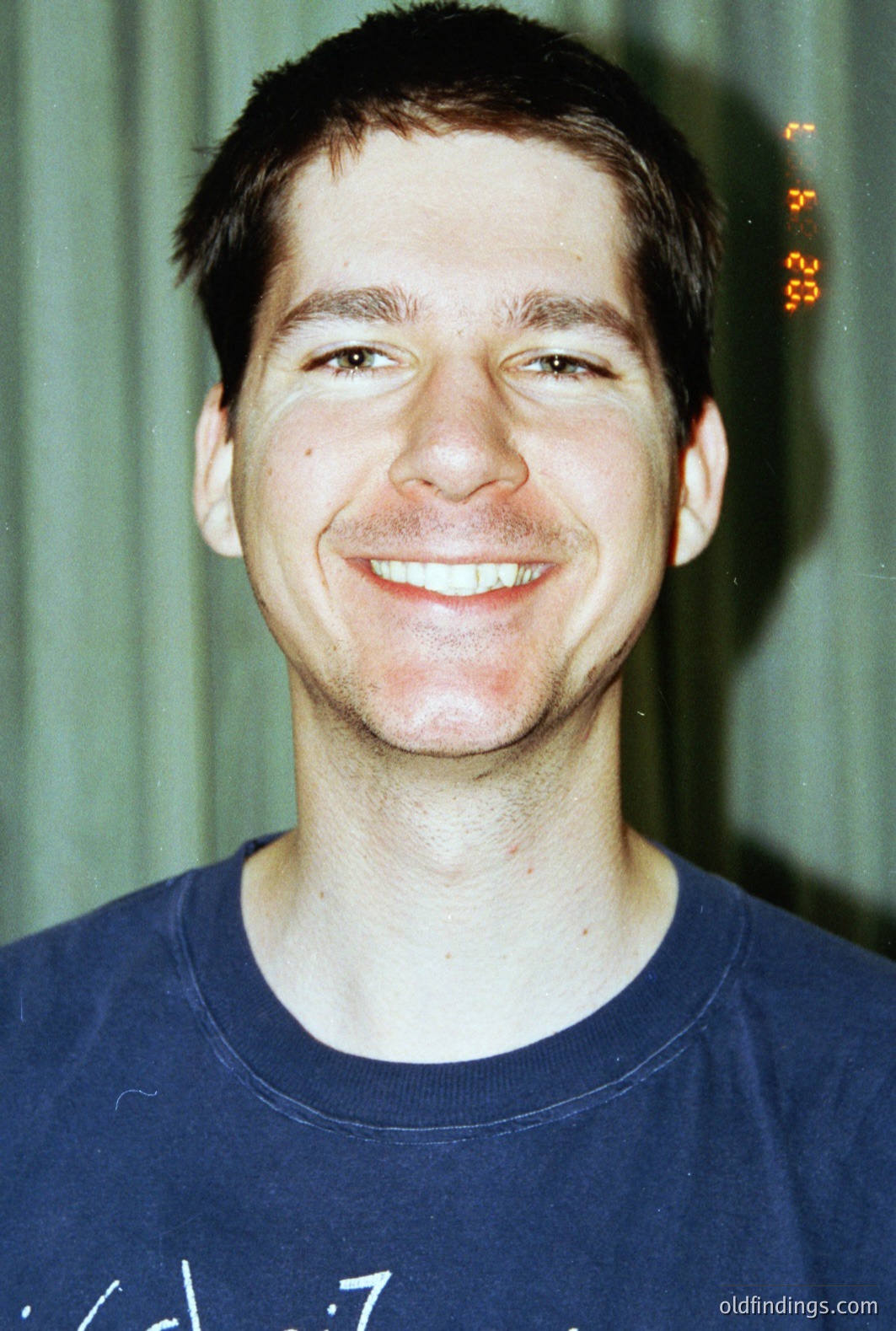 Close-up portrait of a young man with short dark hair, smiling broadly. He wears a blue t-shirt. The background appears to be a green, textured wall. Likely a snapshot from the 1990s given the color palette and film quality. Casual, candid moment.