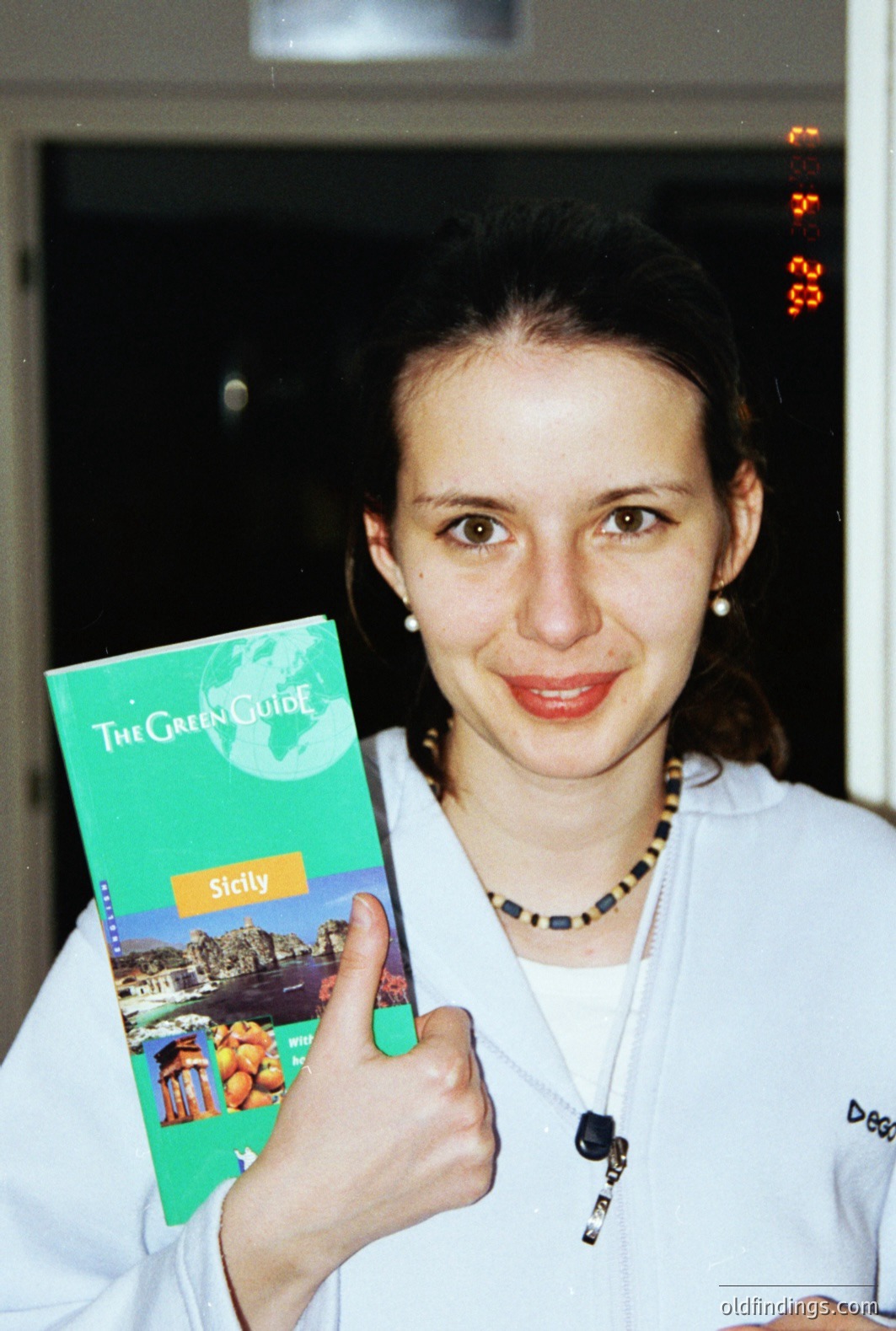 A woman holds a “Green Guide: Sicily” travel book and gives a thumbs-up. She's wearing a white zippered jacket and a dark beaded necklace. The image displays a dated aesthetic, likely a snapshot from the 1990s. The date "08/28/98" appears in the upper right corner. Likely a travel souvenir.