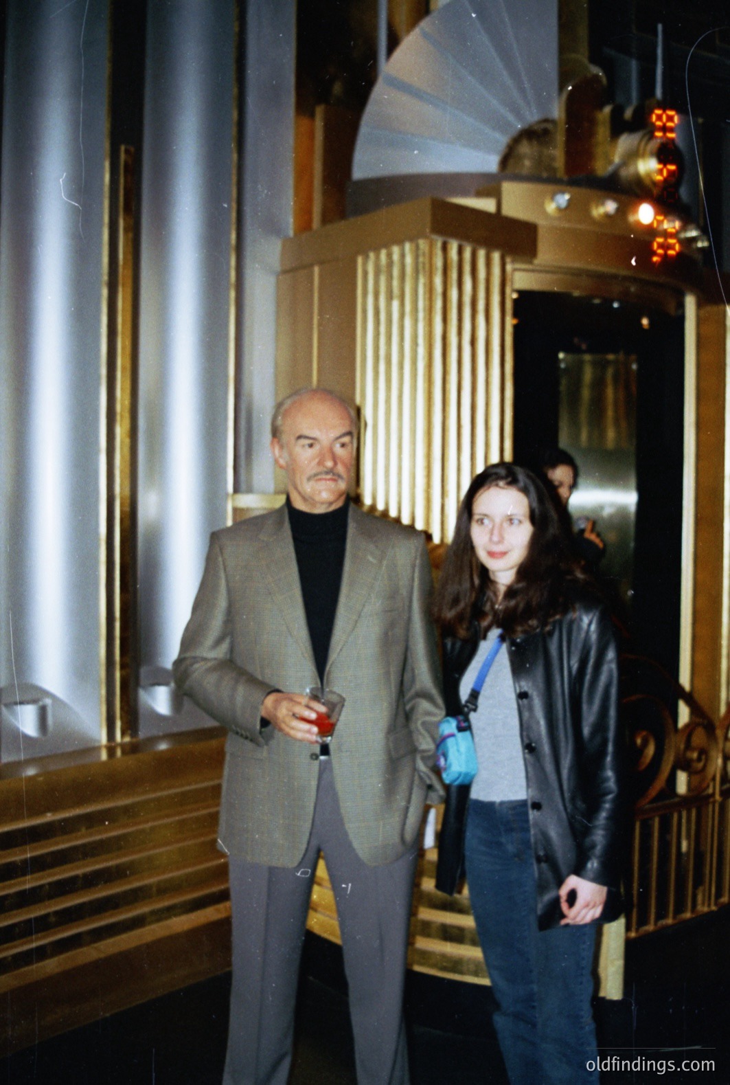 A distinguished gentleman in a tailored suit and turtleneck holds a drink in a grand, gold-accented lobby. A young woman in a leather jacket stands beside him near an elevator. Likely late 1990s or early 2000s, judging by the style. Interior architecture suggests a luxury hotel or apartment building.