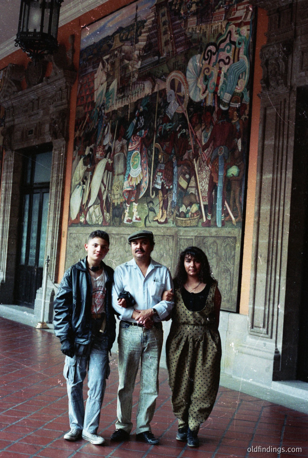 Three figures pose before a vibrant mural in what appears to be a grand, colonial-era courtyard. The mural depicts figures in traditional attire. The subjects—two young people and an older man—are dressed in casual 1980s/early 90s attire. Likely taken in Mexico, showcasing cultural heritage & tourism.
