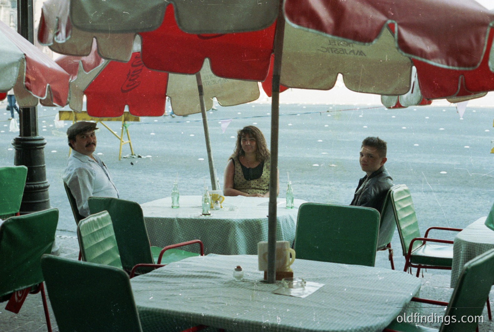 Patio scene: Three individuals sheltered under striped awnings during rainfall. Man in a cap and white shirt stands, while a woman and a man in a leather jacket sit at a table with green linen. Likely a seaside restaurant or café. Appears to be 1970s-80s.