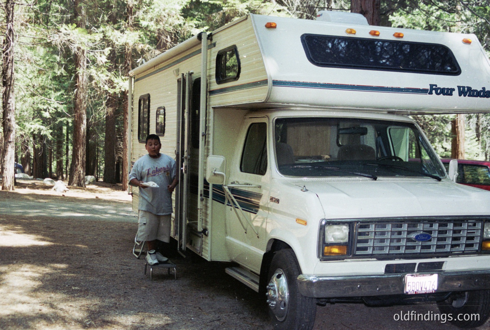 A young boy stands in the doorway of a vintage Ford Econoline campervan, holding a paper plate. The vehicle features “Four Winds” branding and distinctive 1970s-80s styling. Situated in a wooded campsite, pine trees dominate the background. Likely a family vacation photo.