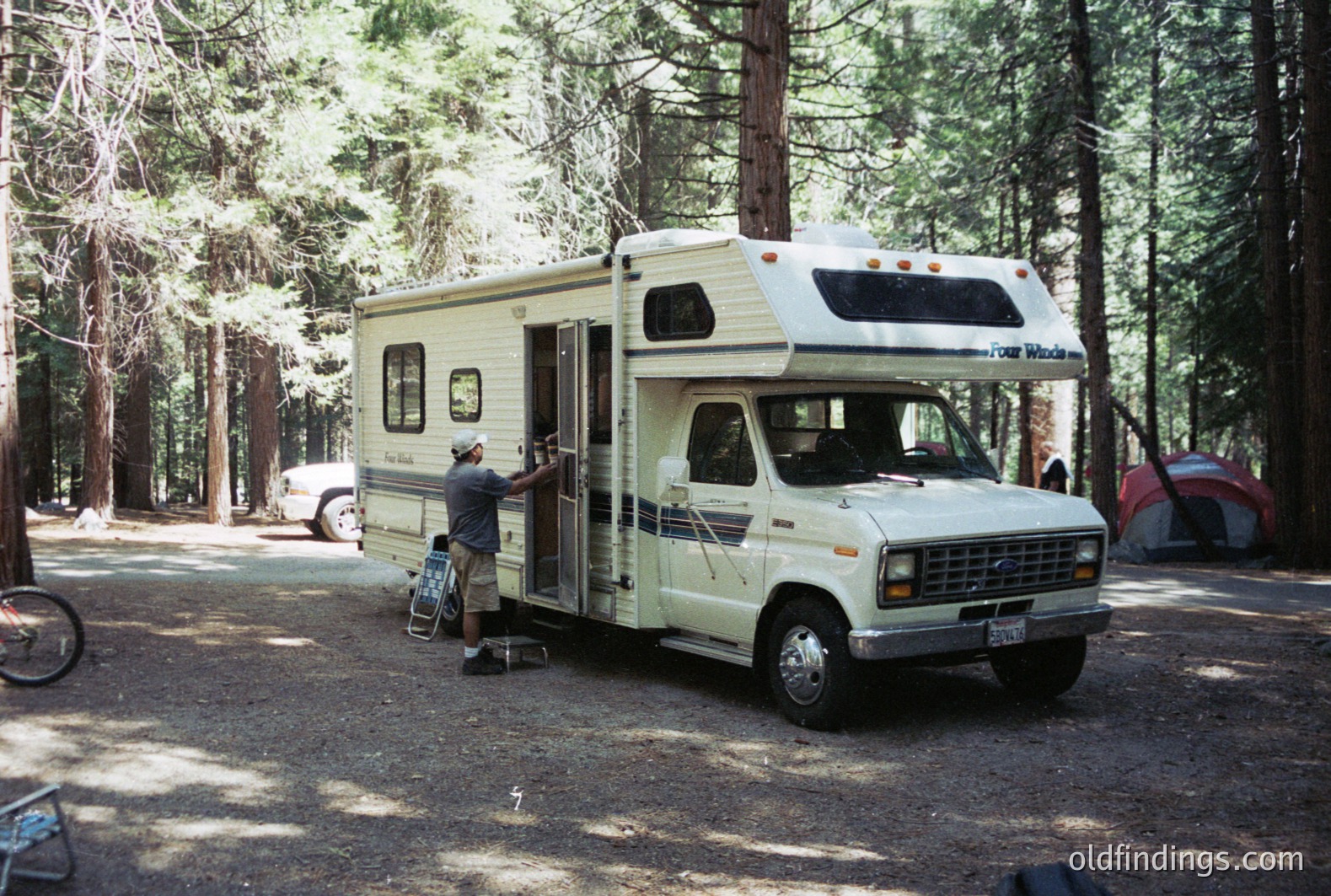 A vintage "Four Winds" Ford Econoline conversion van, likely from the 1980s, is parked in a forested campground. A man, wearing a hat and shorts, is standing next to the open door. A bicycle leans nearby, with other campsites visible in the background. Scenic and nostalgic travel imagery.