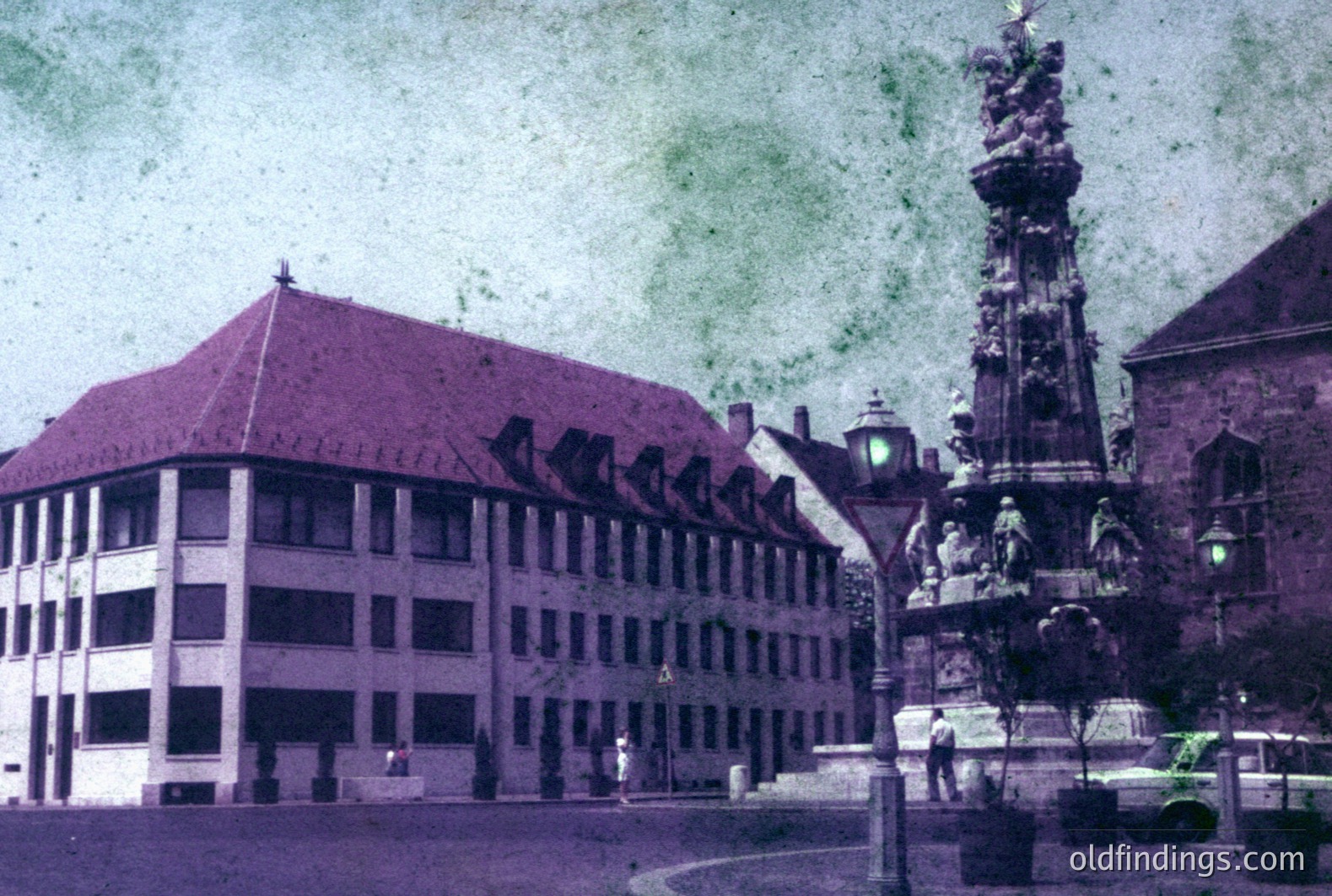 A vibrant, early-color photograph depicts a classical fountain monument & adjacent building with a red-tiled roof. The building's facade features numerous rectangular windows. A lone figure is seen near the fountain. Appears to be a European city square, likely 1950s or 1960s.