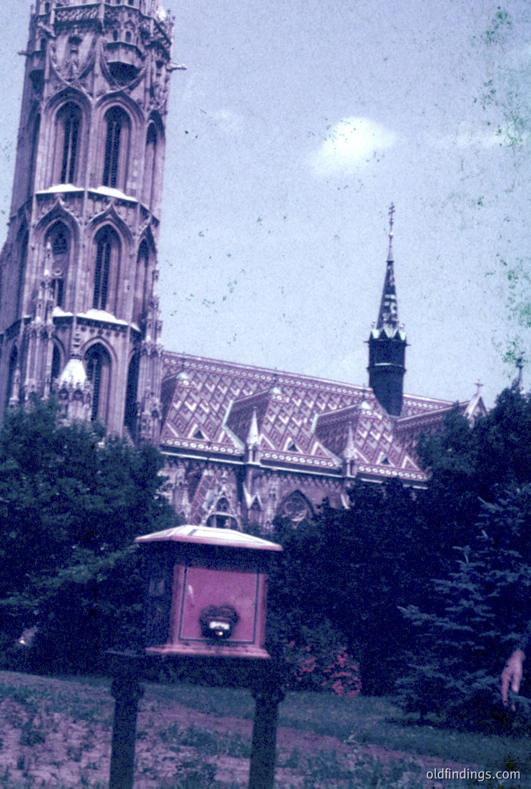 An imposing Gothic Revival church dominates the view, featuring a prominent spire and intricate detailing. A small, ornate pavilion stands in the foreground. Likely a commemorative structure, it hints at a historical or memorial context. The image suggests a mid-20th century (1950s-1970s) aesthetic with faded colors.