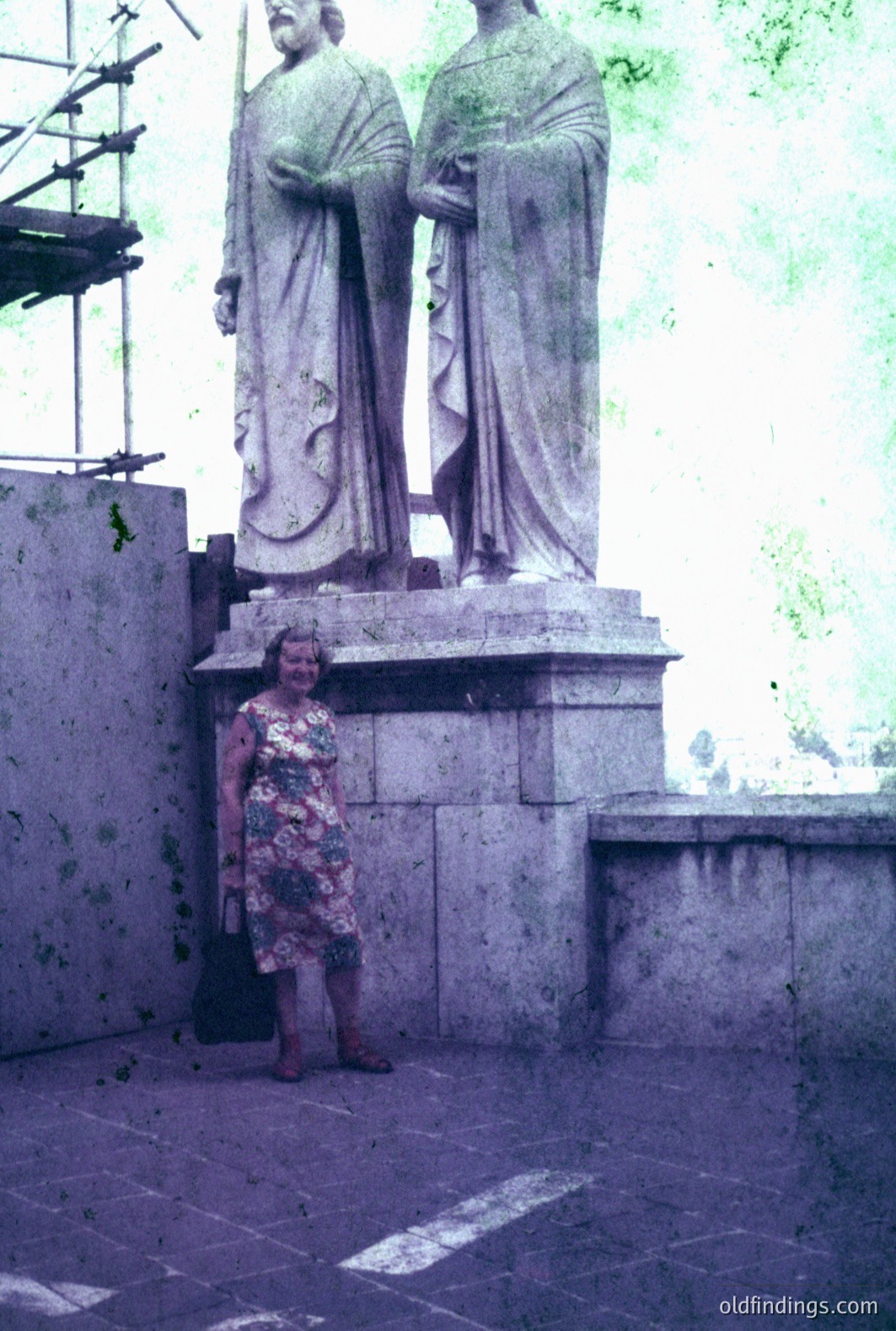 A woman stands near a large, classical-style stone statue of two figures, likely commemorating historical figures. She wears a floral-patterned dress and sandals. The setting appears to be a courtyard or plaza with visible foliage. The photograph’s faded colors and visible film grain suggest a date in the 1970s or 1980s. Potential stock photography reference for travel or historical tourism.