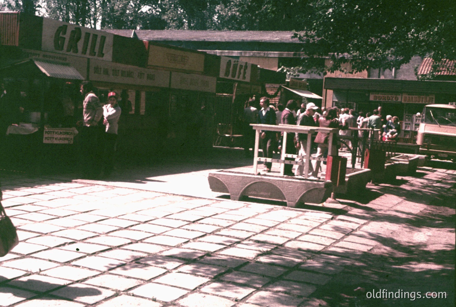 A low-angle view captures a vintage miniature train moving across a tiled plaza. Buildings with signage "Grill" and "Buffet" line the perimeter. Several people are on and around the train. Likely a park or seaside attraction, 1960s-1970s. Location unknown.