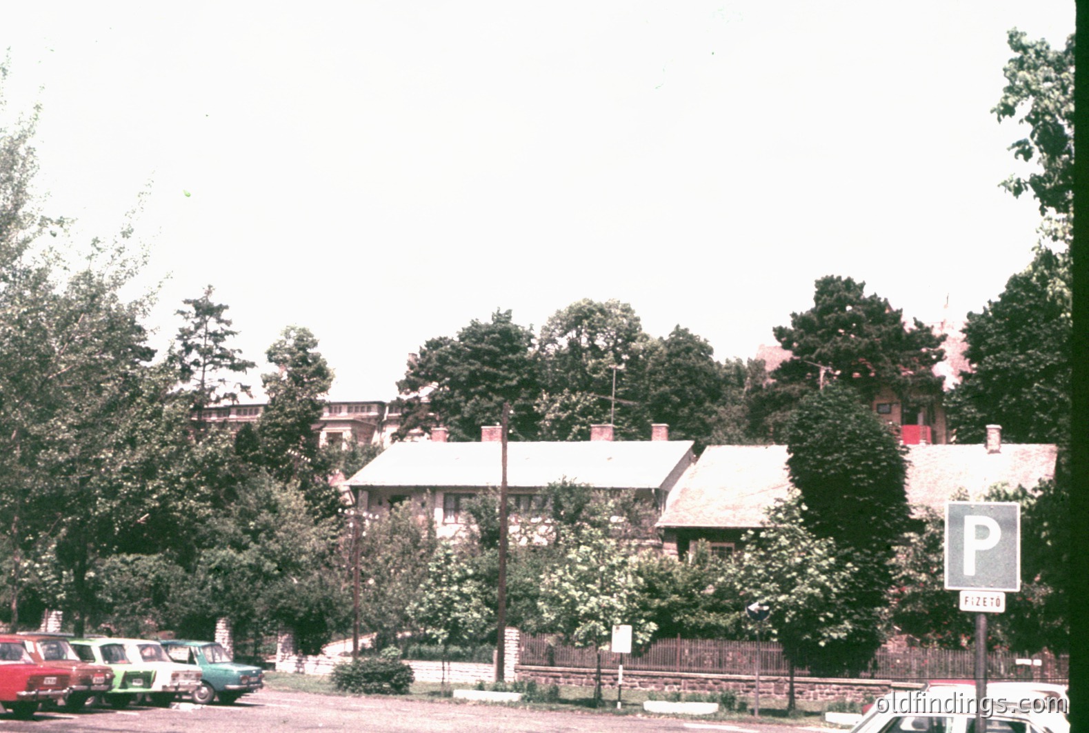 A roadside view showcases traditional, low-rise buildings with tiled roofs, likely residential. Lush trees obscure much of the structures. A parking sign indicates a public area. Car models suggest a 1960s-70s timeframe. Potential location: Eastern Europe.