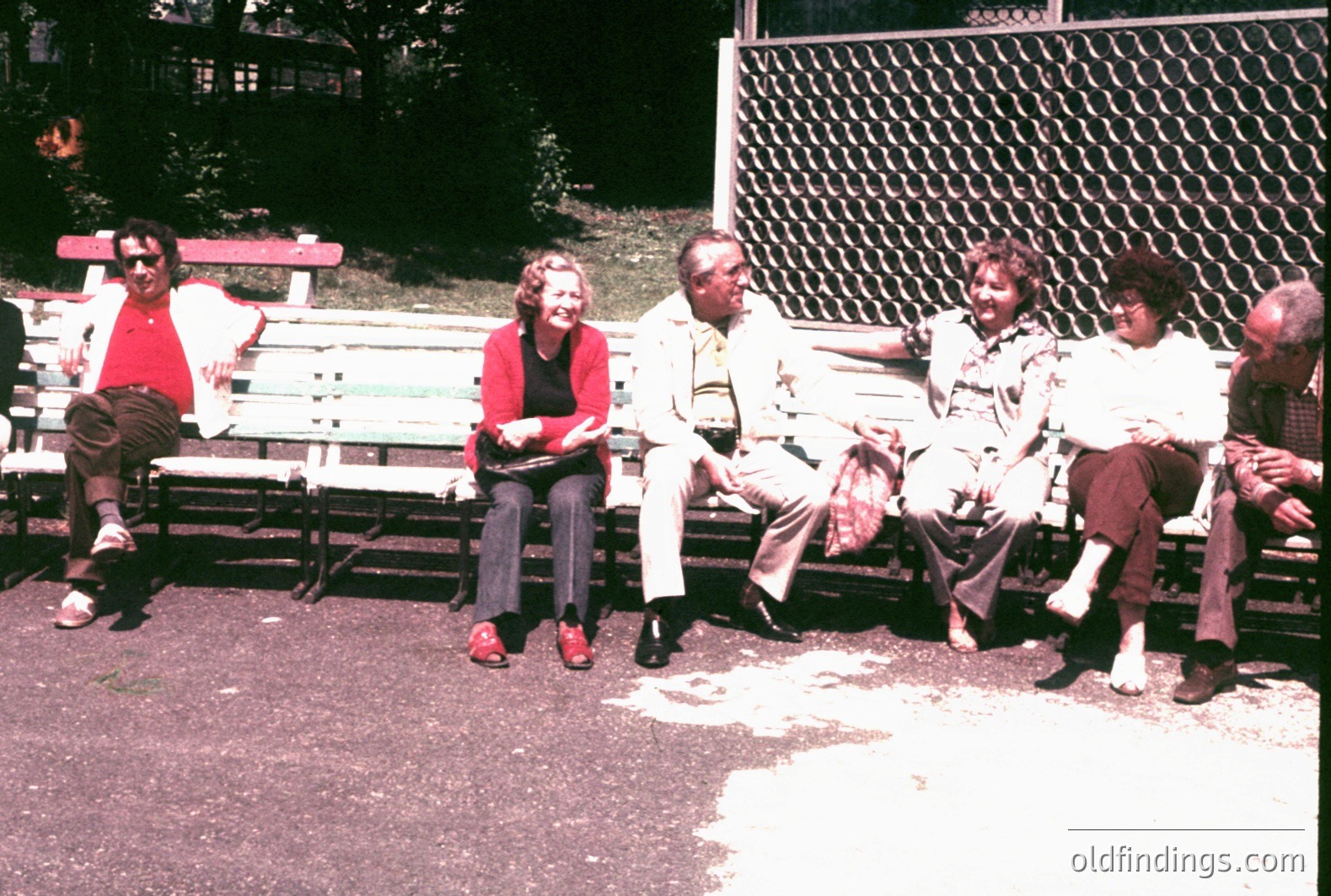 A group of five individuals are seated on a public park bench in what appears to be a casual, relaxed setting. The style of clothing and hairstyles suggest a mid-1970s timeframe. The bench is constructed with a wooden slat design. A chain-link fence with foliage is visible in the background.