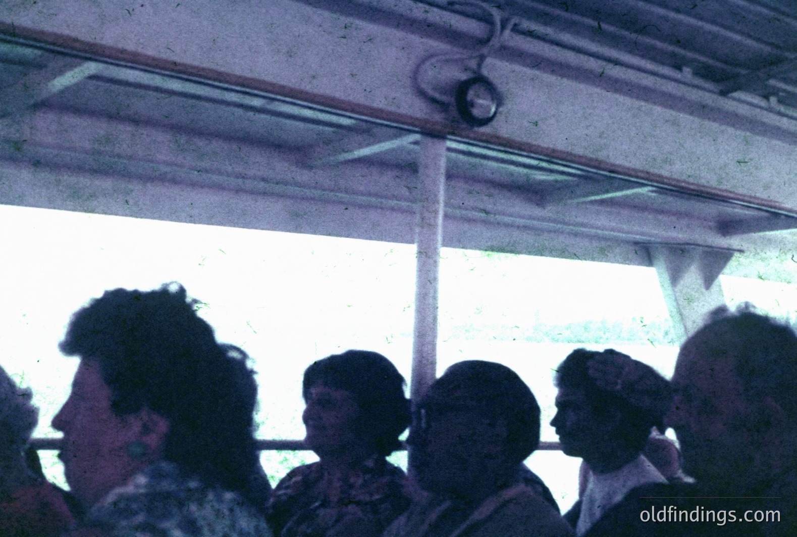 Four individuals are seen through a window, likely on public transportation. The image exhibits a grainy quality characteristic of mid-century film. Clothing styles suggest a 1960s or 70s timeframe. The visible ceiling fixture and window frame contribute to the scene's sense of place.